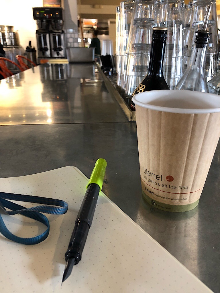 Pen, notebook, and paper coffee cup on a steel restaurant counter.  