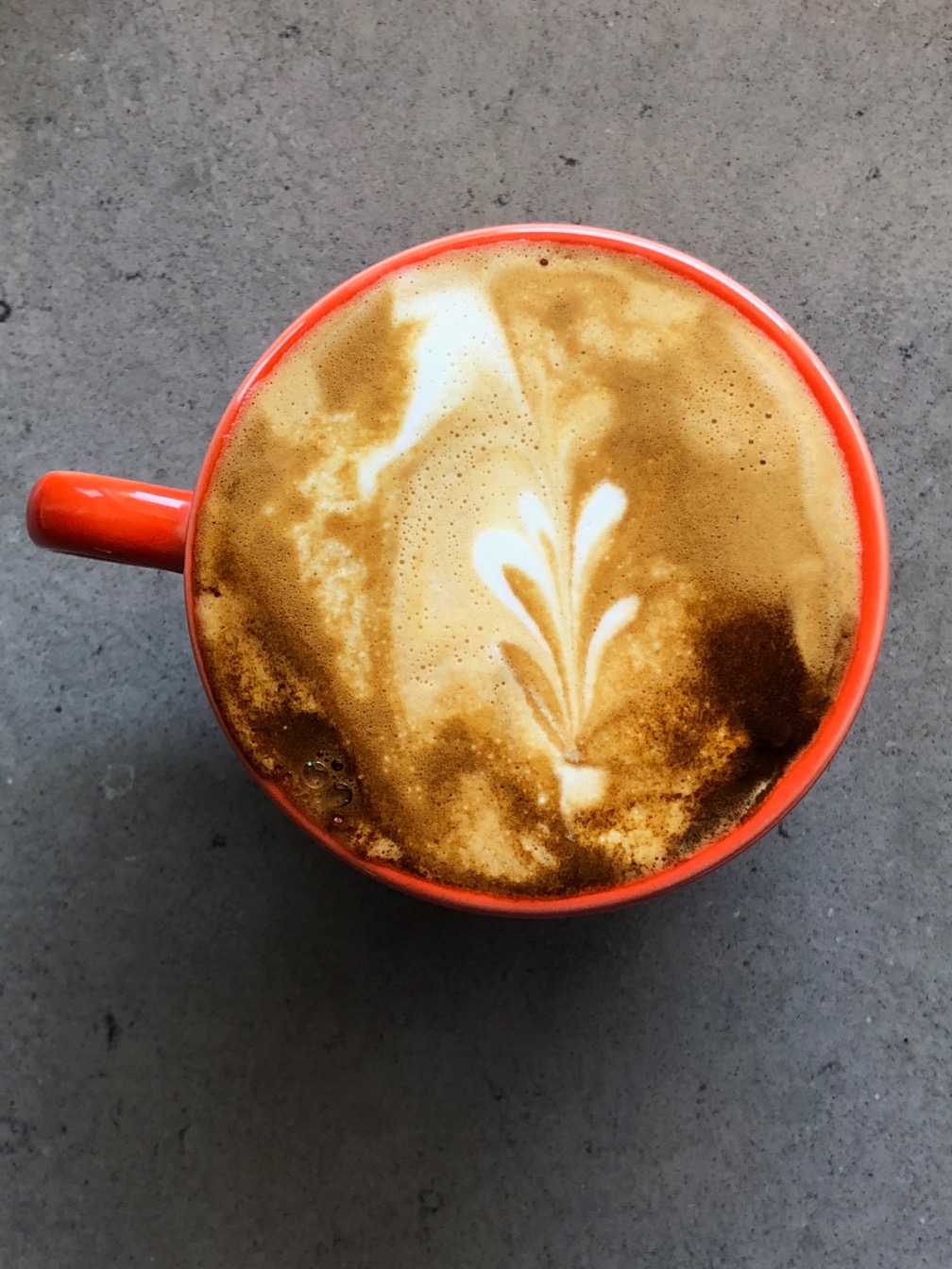 Cappuccino in an orange cup on a speckled gray counter.  