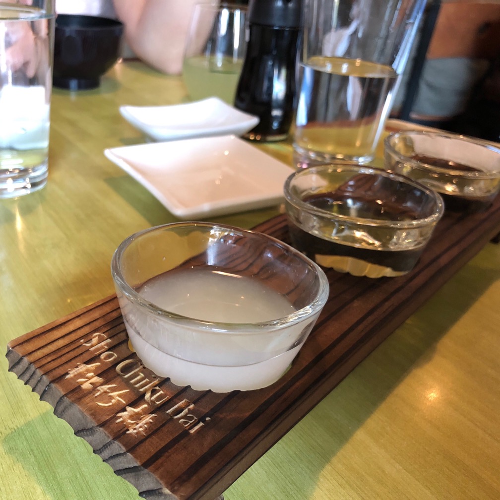 Three small cups of sake on a wooden tray. The front one is cloudy white from unfiltered rice.  