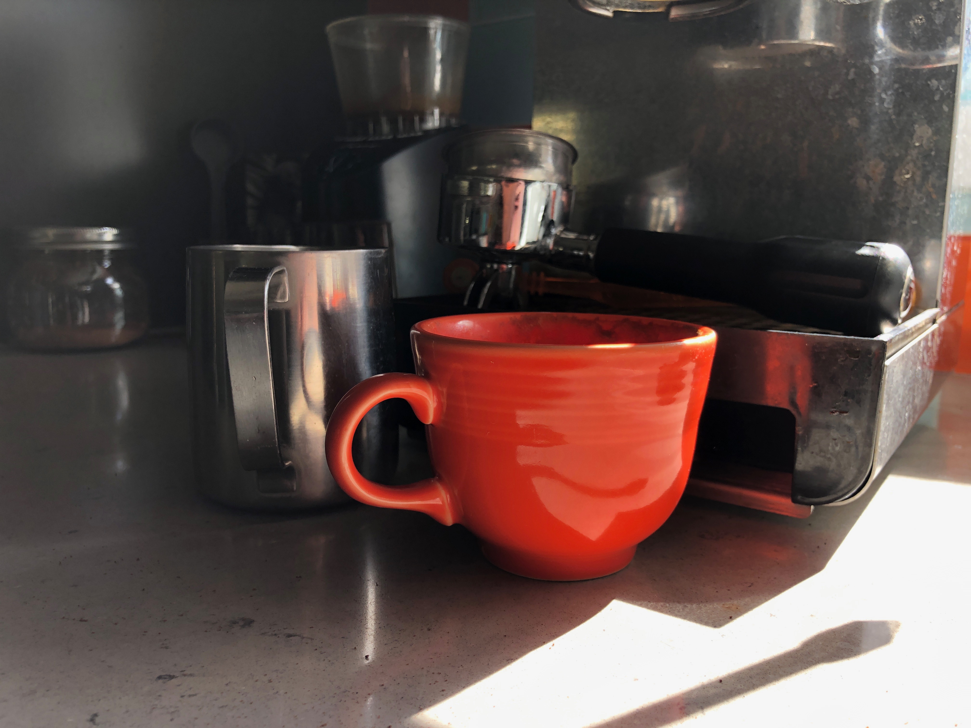 A bright orange mug in front of an espresso machine, portafilter, and milk pitcher 