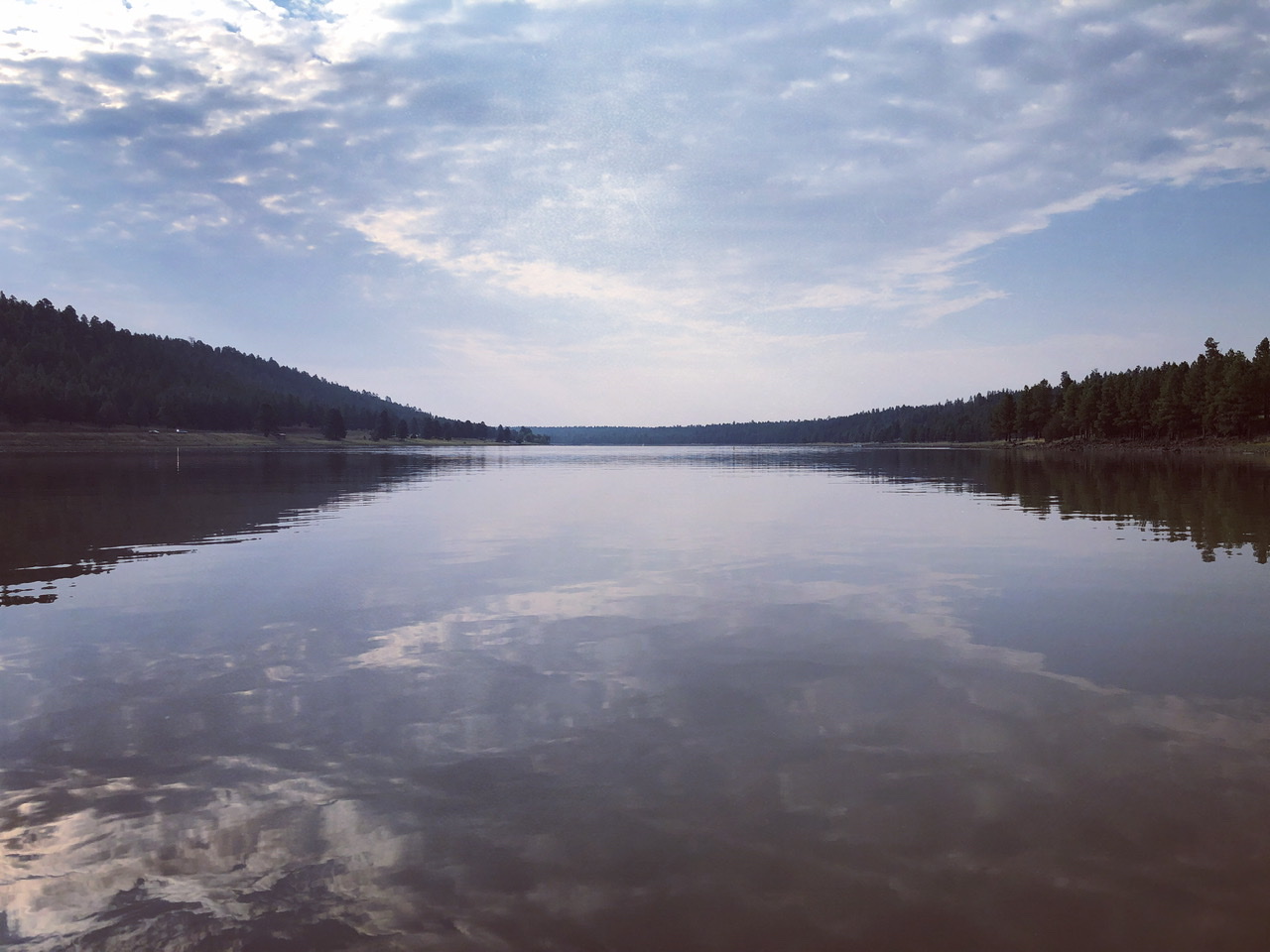 A view across a lake, with clouds and trees reflected in the water 