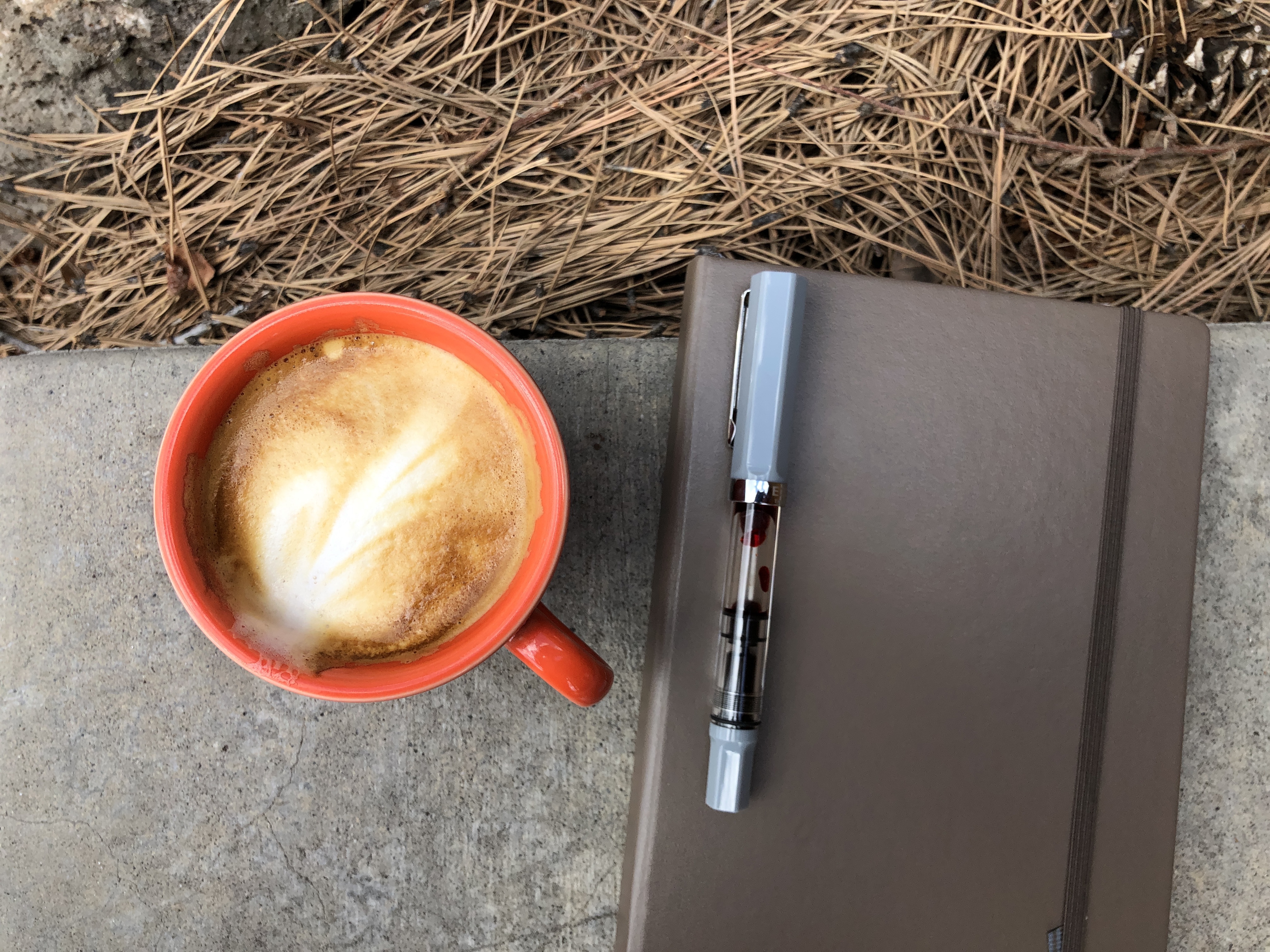 an orange cappuccino mug on a concrete step, beside a notebook and a grey and orange pen 