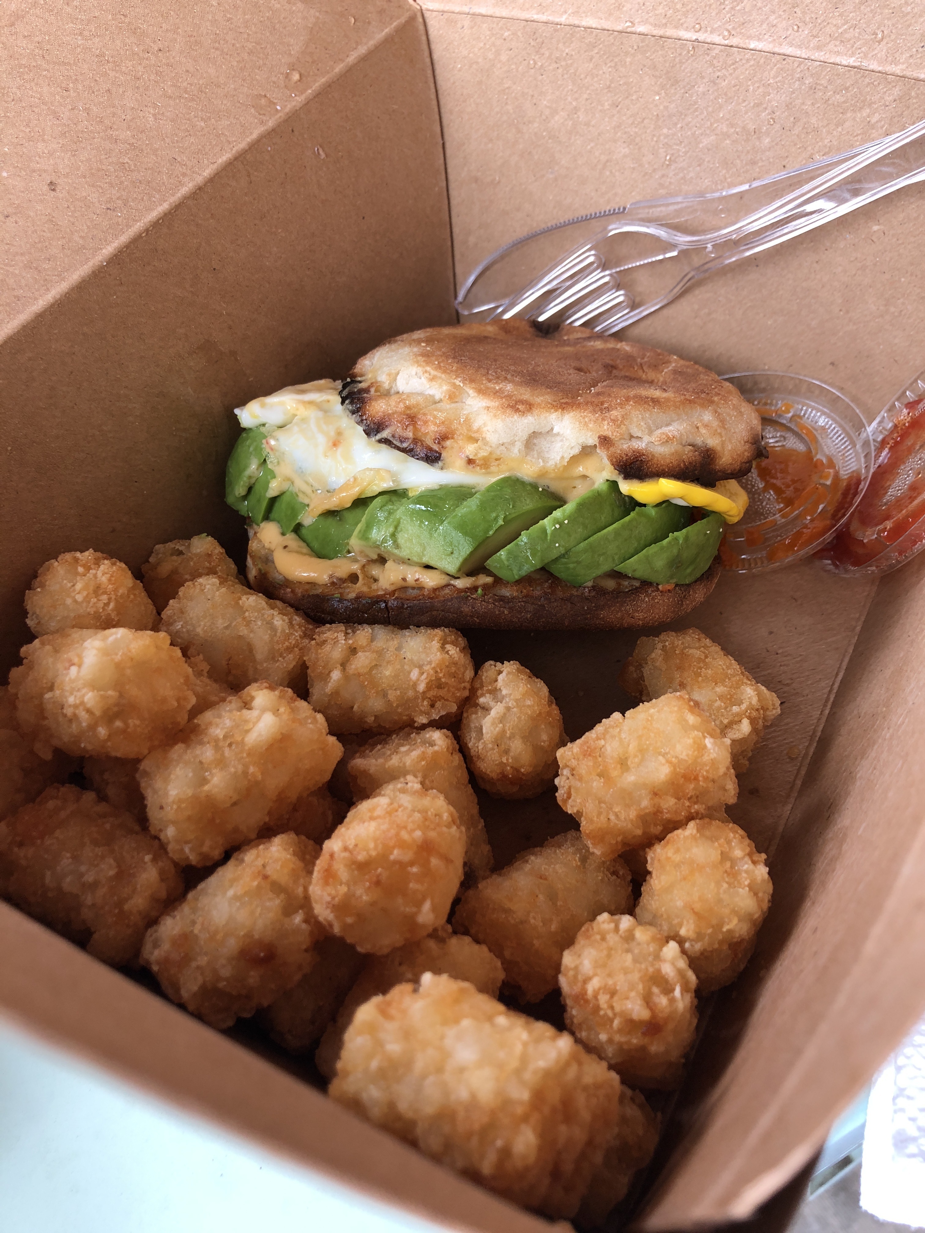 A mound of tater tots surround a breakfast sandwich of avocado and egg on a toasted english muffin 