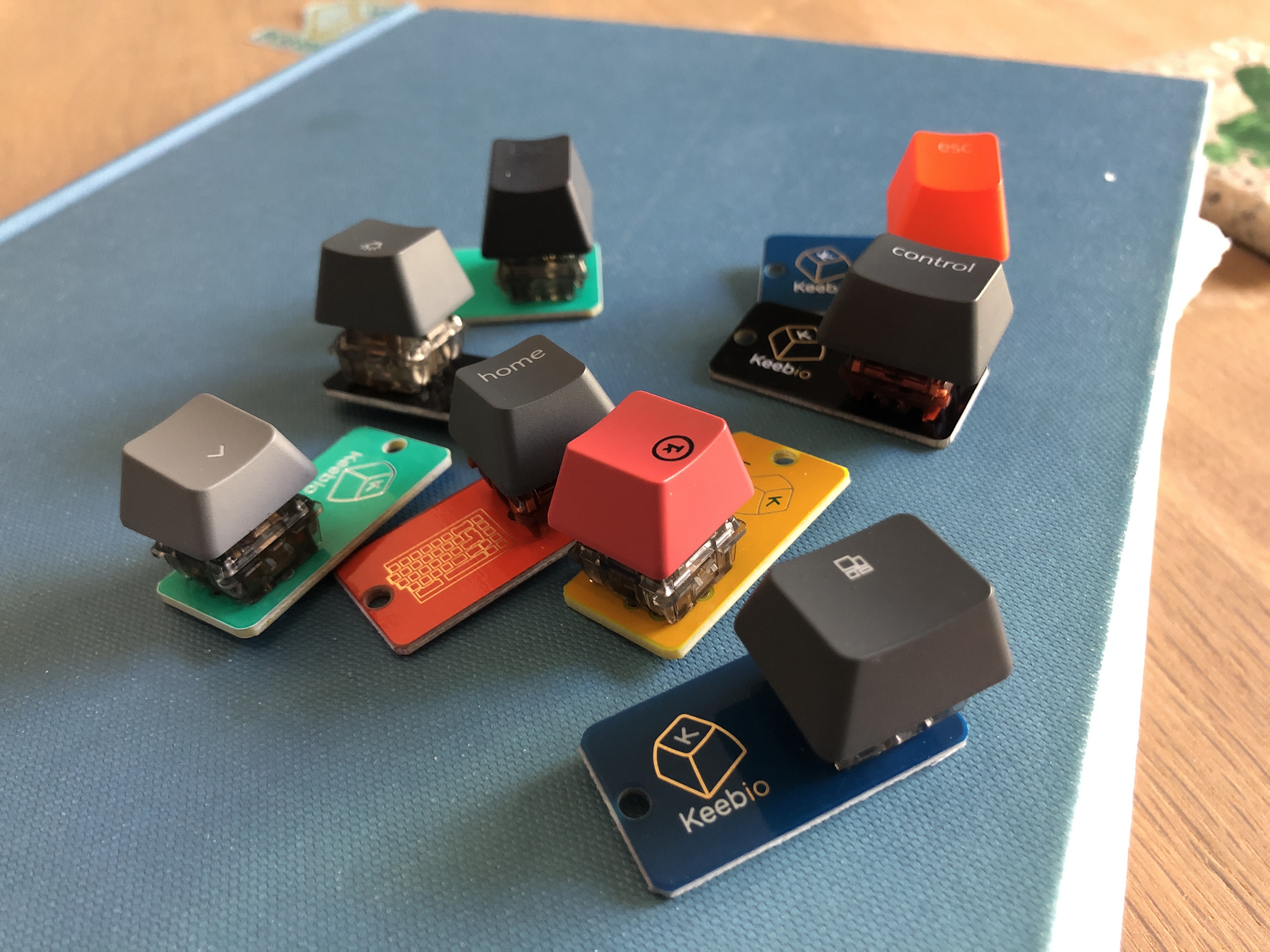 several tiny metal tags in blue, teal and black, each with a single mechanical keyboard switch and variety of keycaps mounted on top. They are arranged haphazardly on a book. 