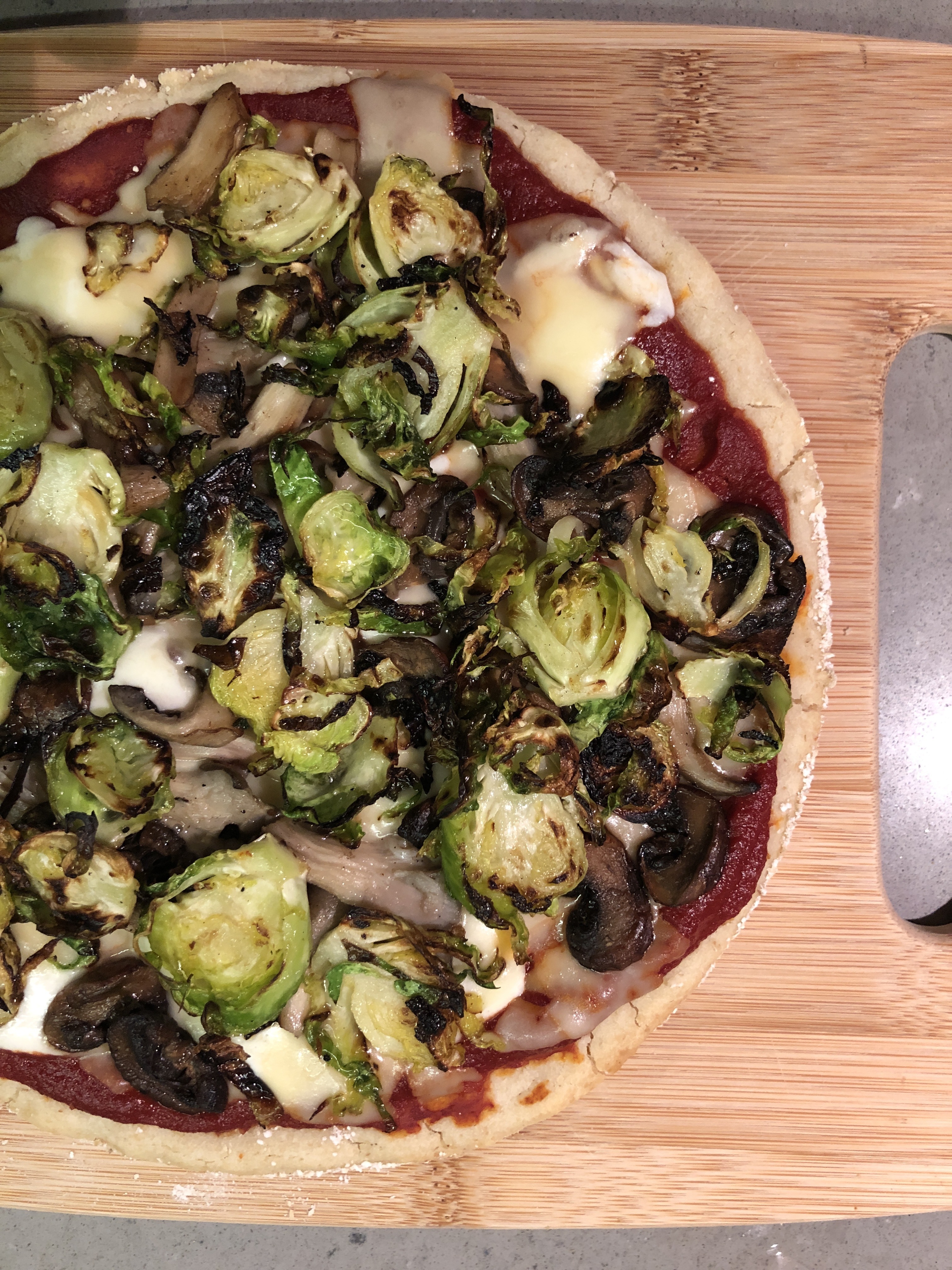 a pizza sitting on a wooden cutting board. It's topped with bright green slices of cooked sprouts, contrasting with white cheese and red sauce. 