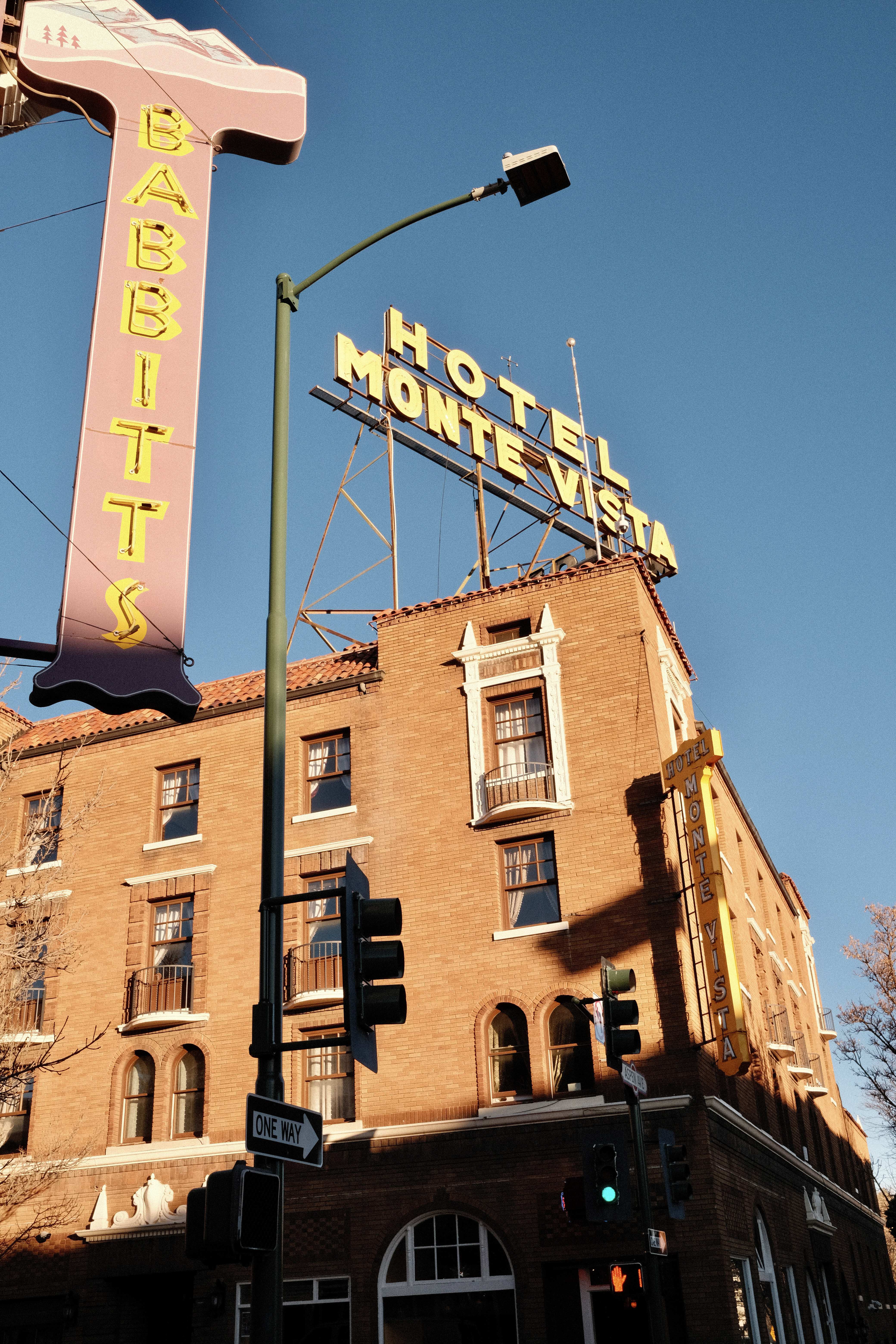 A brick building on a corner with a large yellow sign for the Hotel Monte Vista above it, lit by late evening sun 