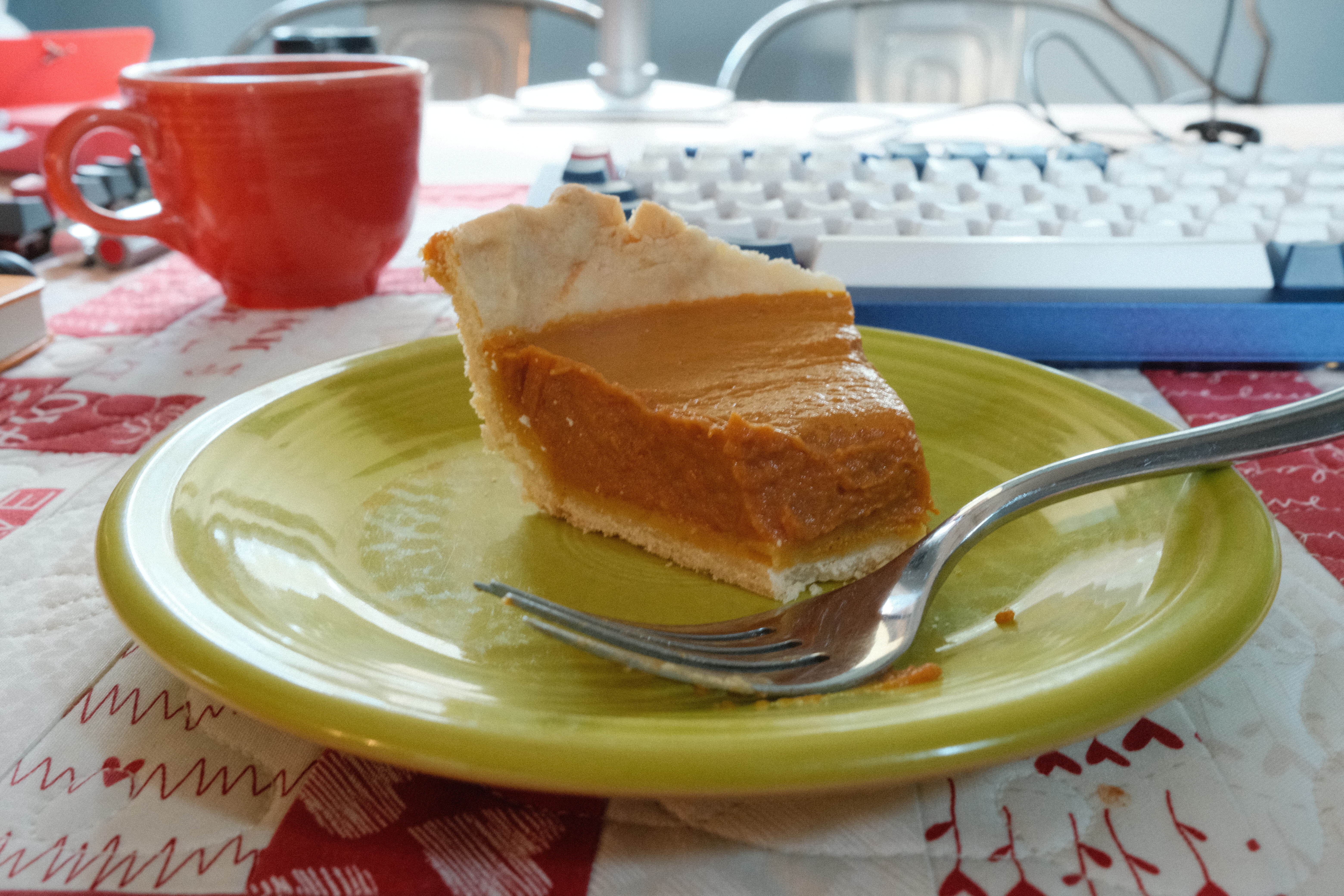 a slice of pumpkin pie on a bright green plate, in front of a white-keyed keyboard and orange coffee cup 