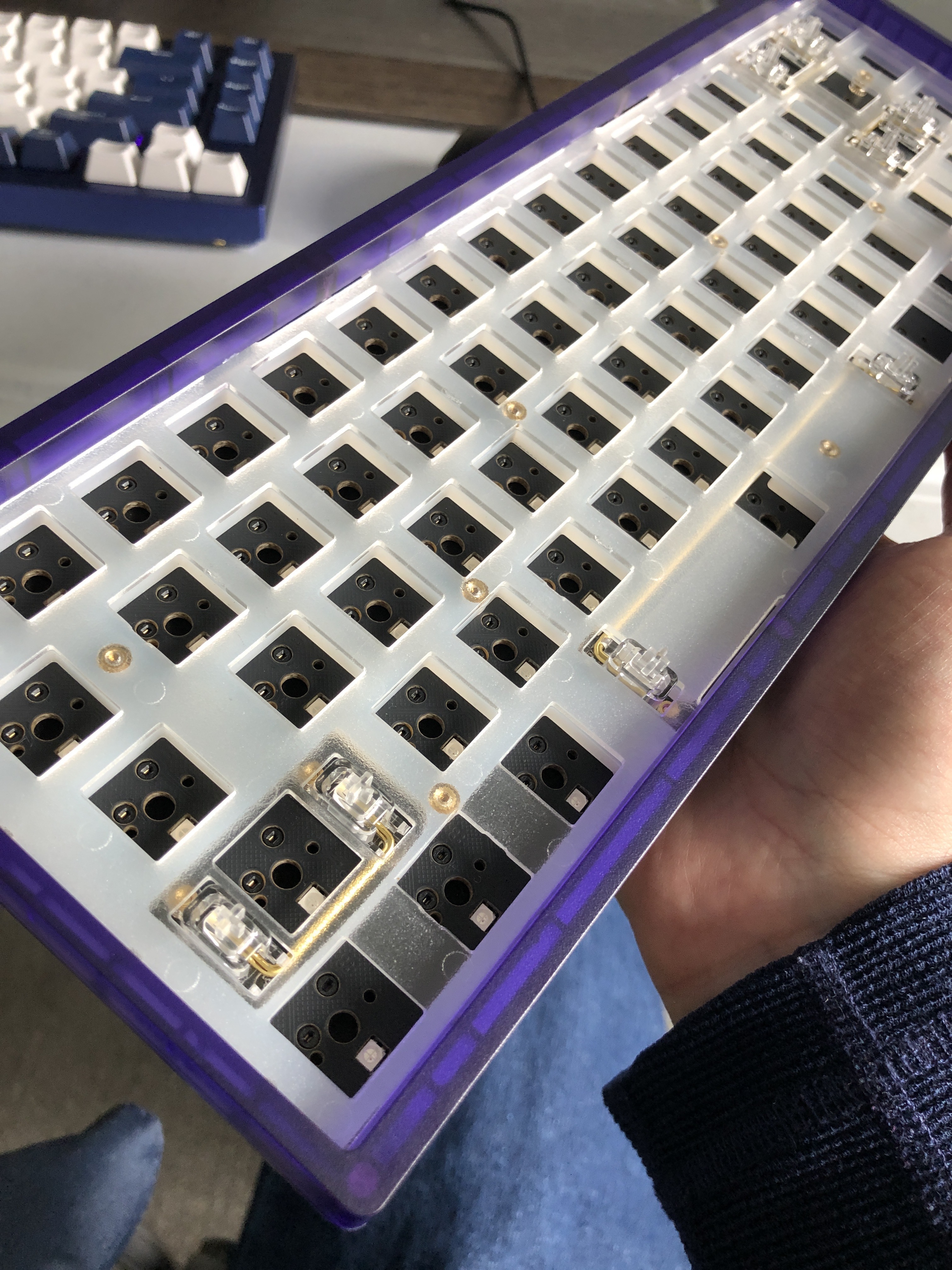 A blue plastic keyboard case without switches or caps installed, showing the bare switch sockets, held in my hand 