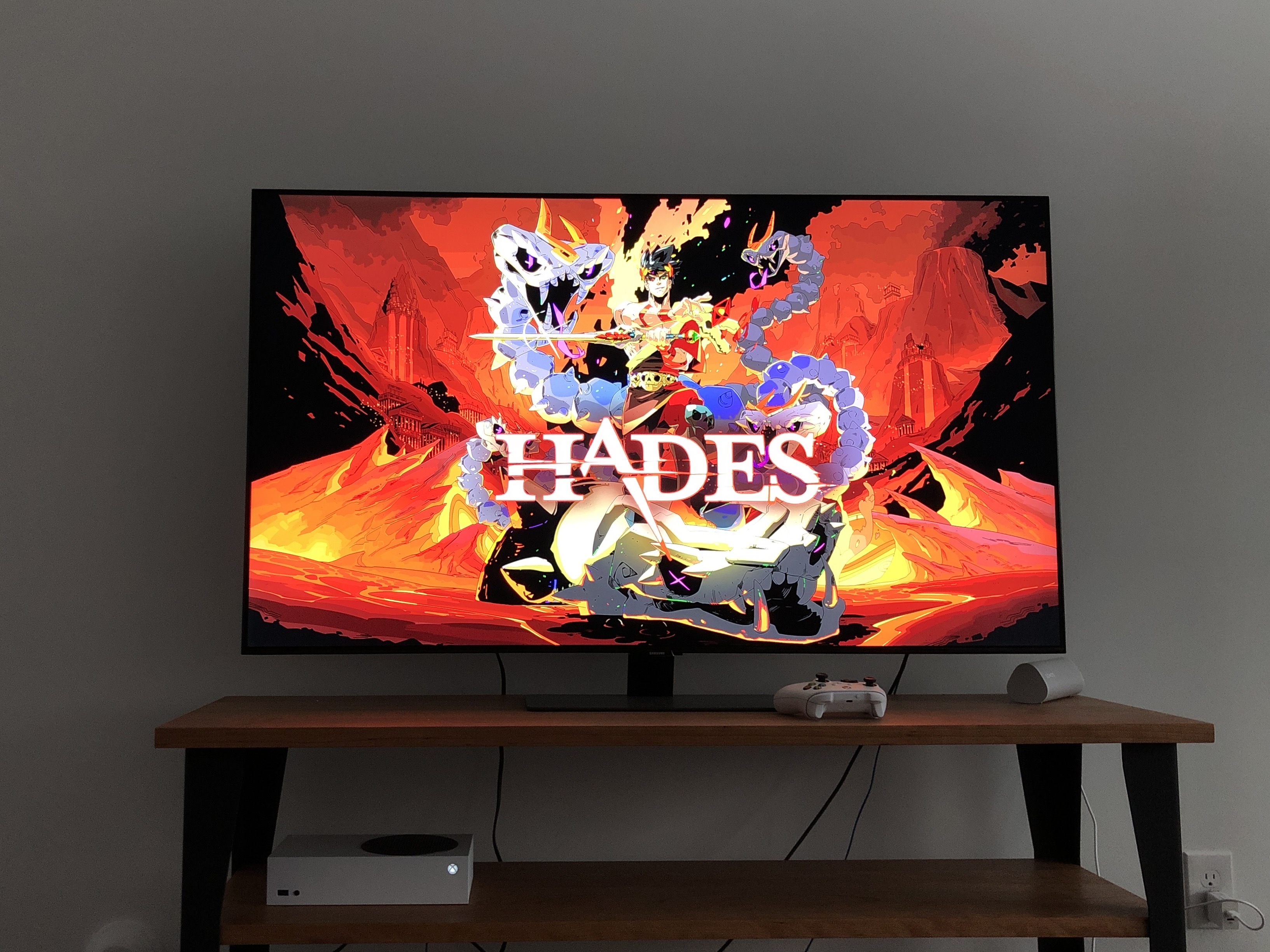 A TV showing the title screen for Hades, with an Xbox Series S and white controller in the frame, also 