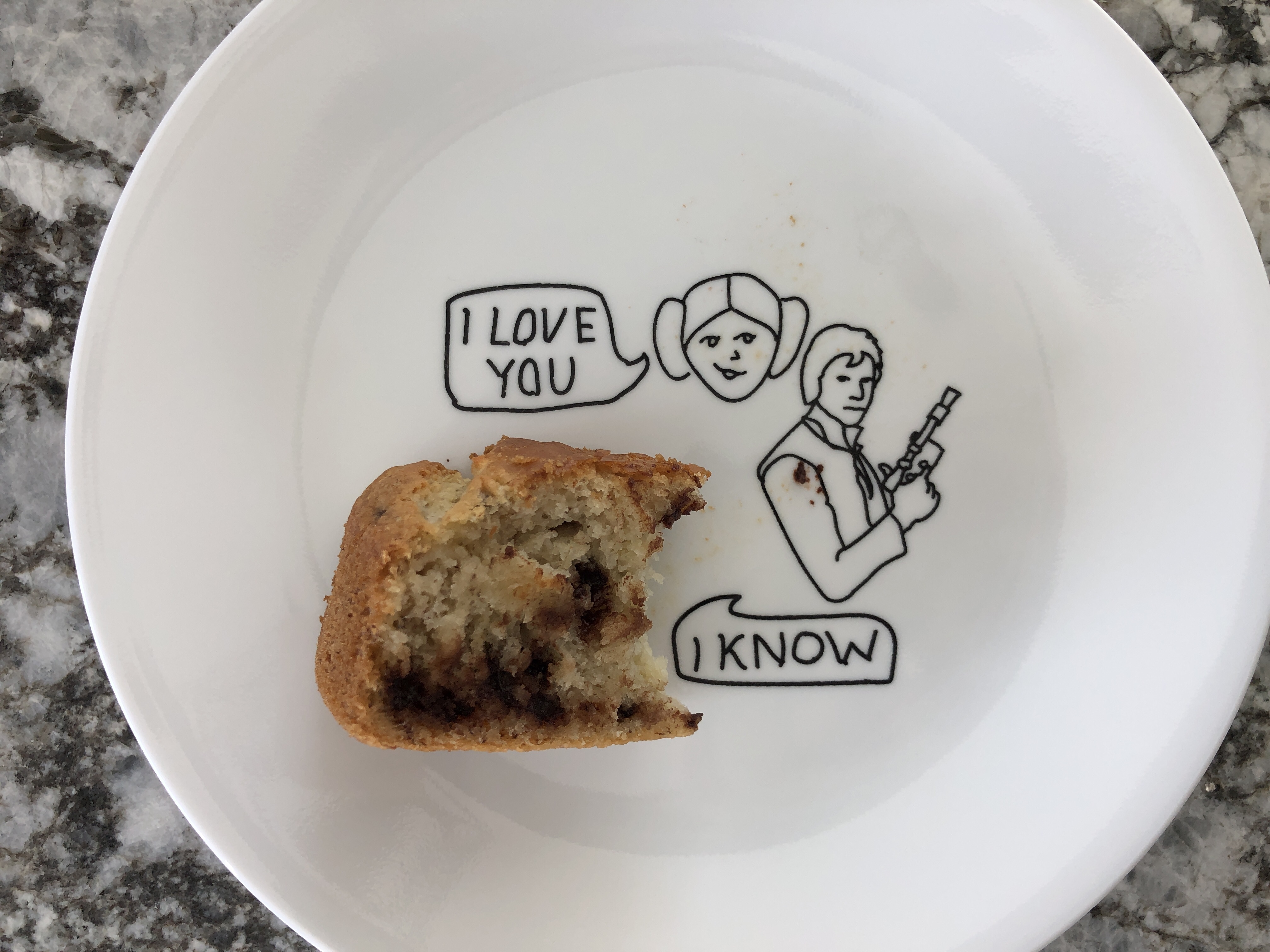 A half eaten slice of banana bread sits on a plate on which is drawn a cartoon figure of Leia saying ‘I love you’ to Han saying ‘I know’.