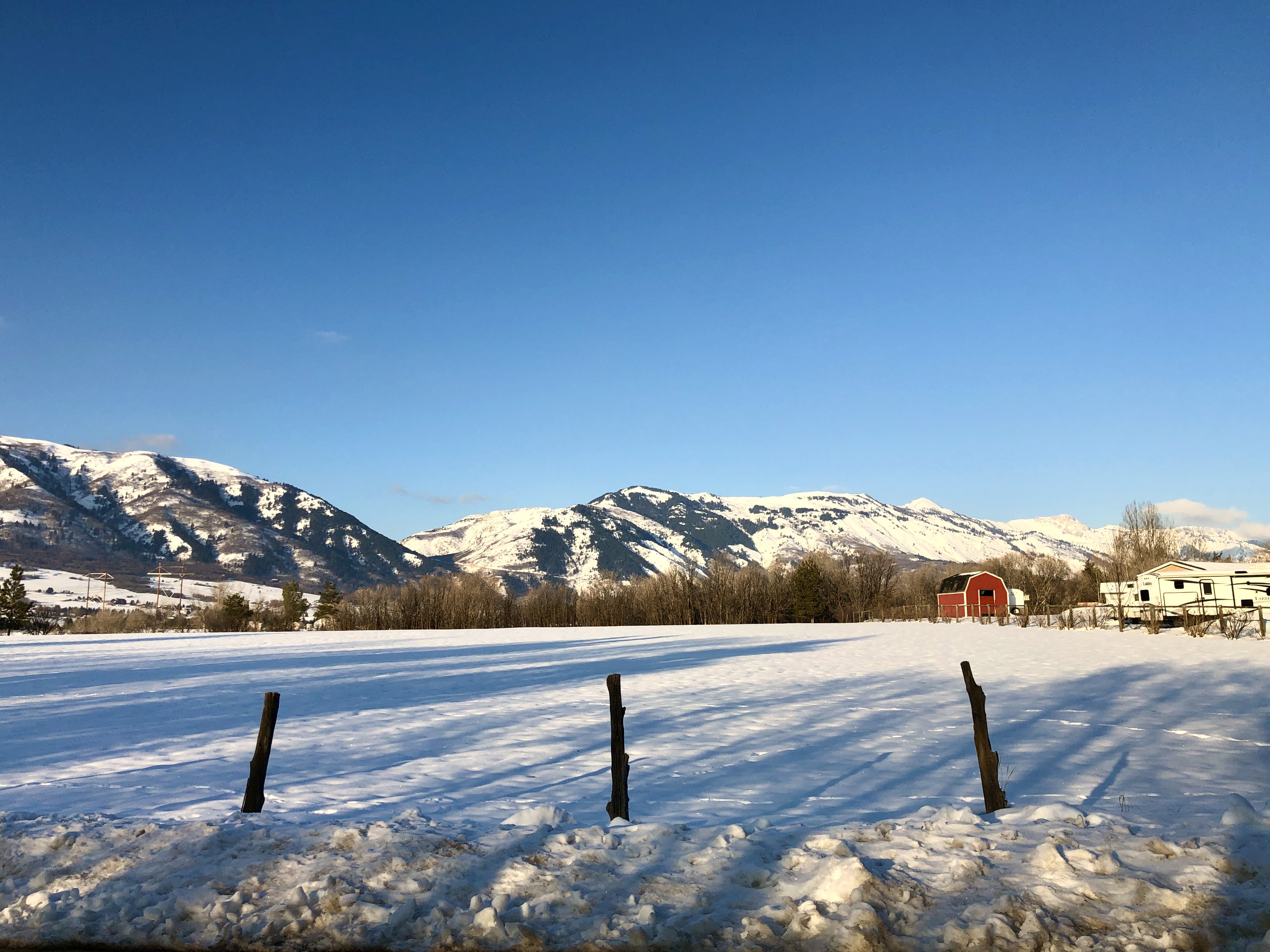 A snowy field lies beyond a set of broken fenceposts, with a red barn at one side. Behind it is a ridge of mountain and bright blue sky is above.