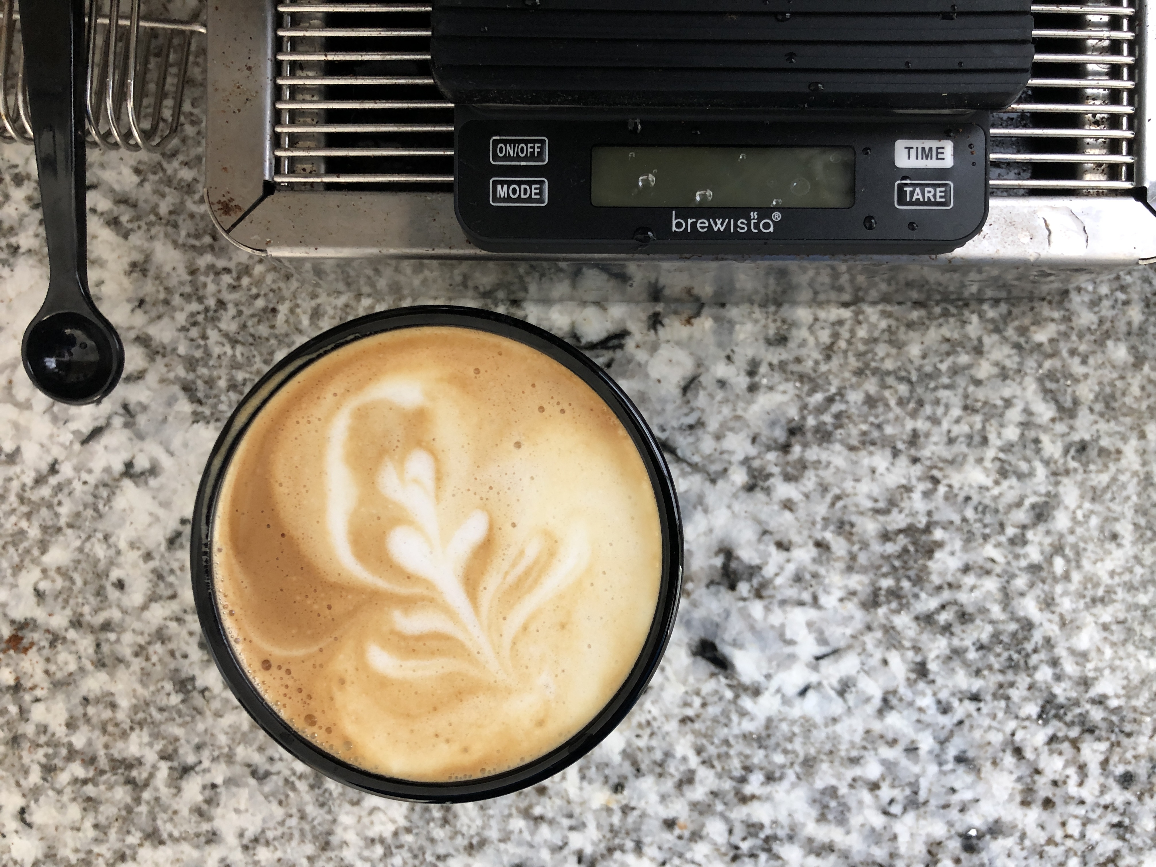 a cappuccino in a gray cup on a black and white speckled countertop, beside a small digital scale