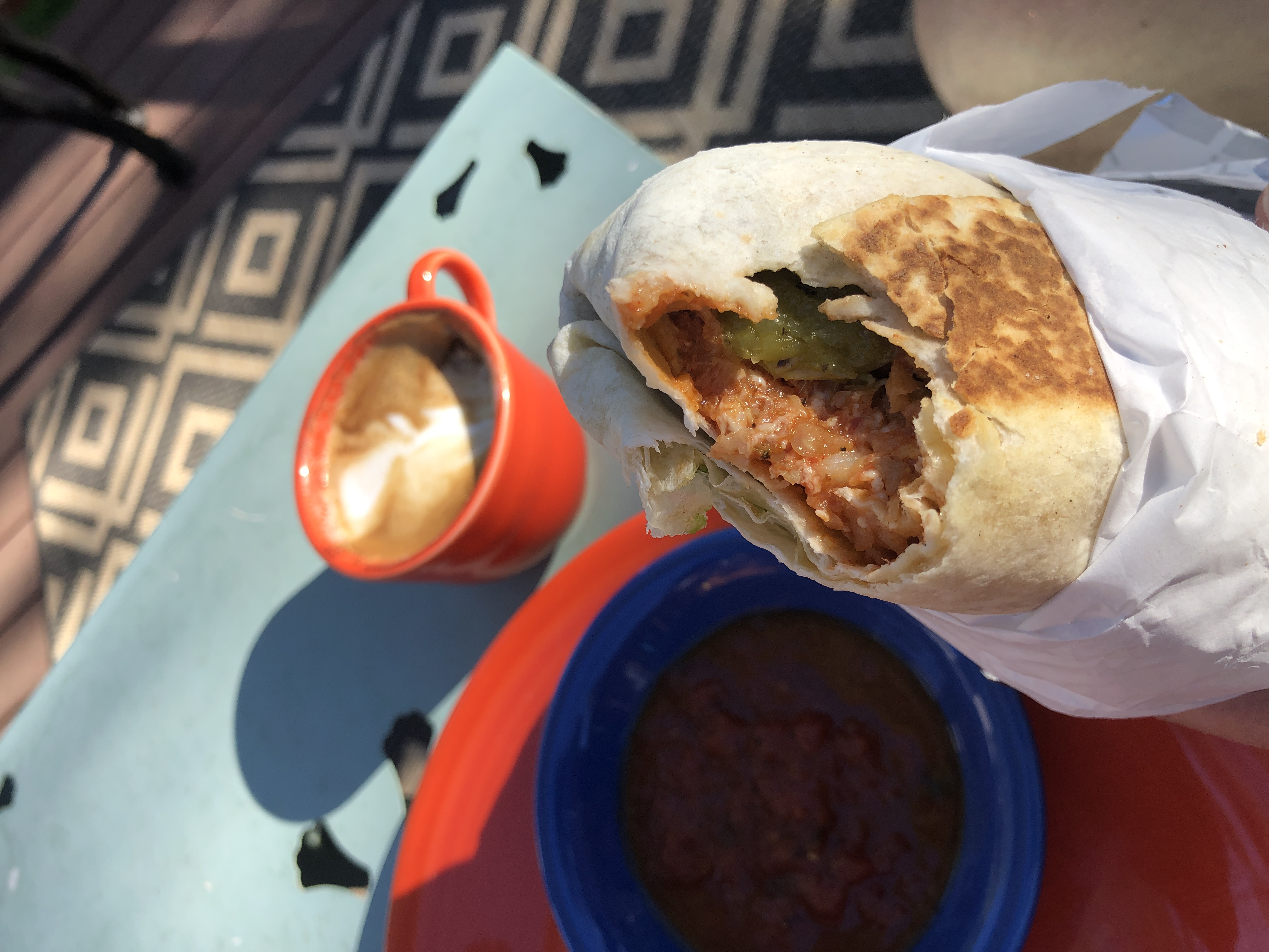 A large burrito with one bite taken out, with a coffee cup in the sun behind it