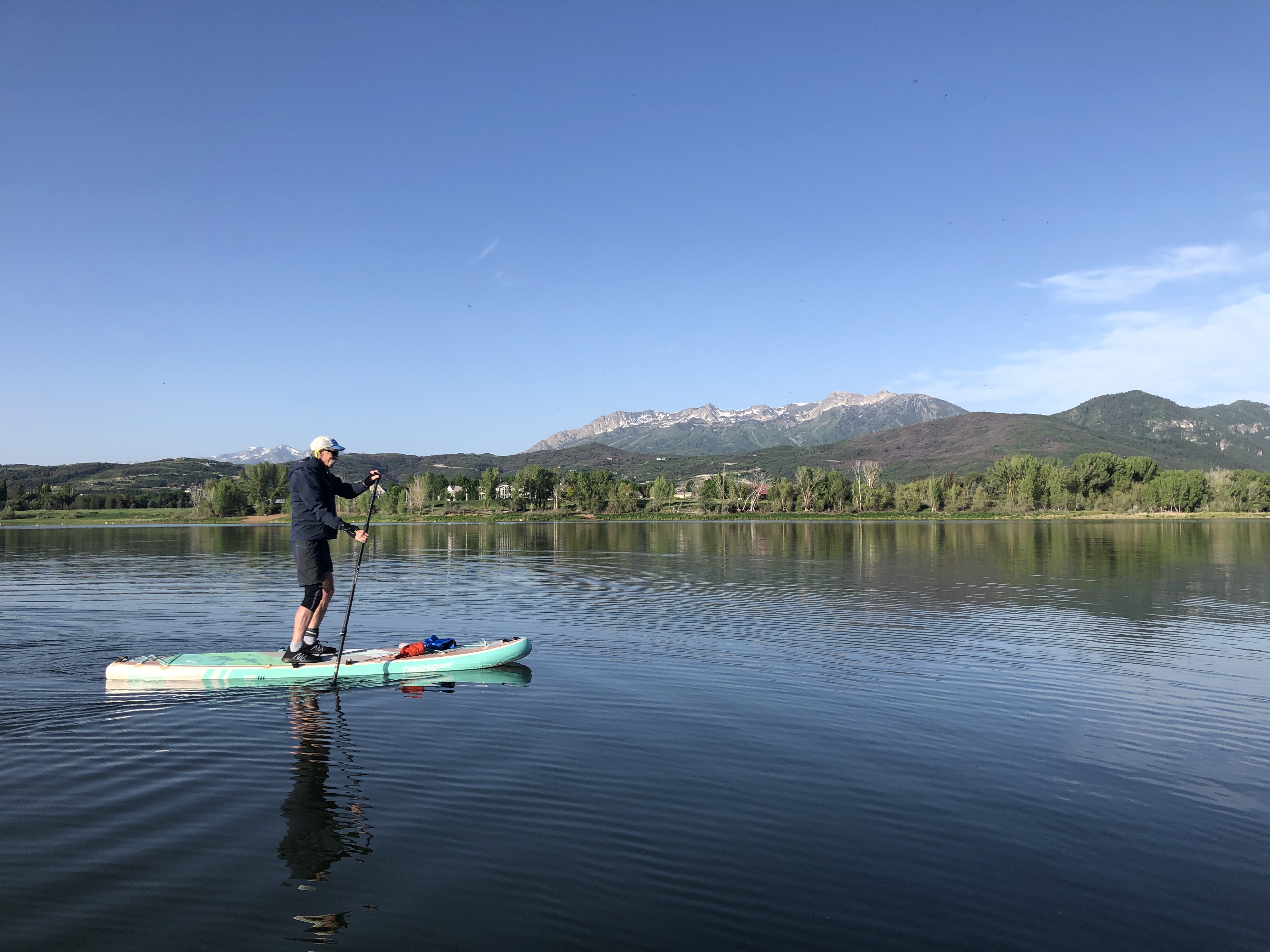 A man standing on a light blue paddleboard, with water stretching to trees and then a background of peaks with a bit of snow still on them.