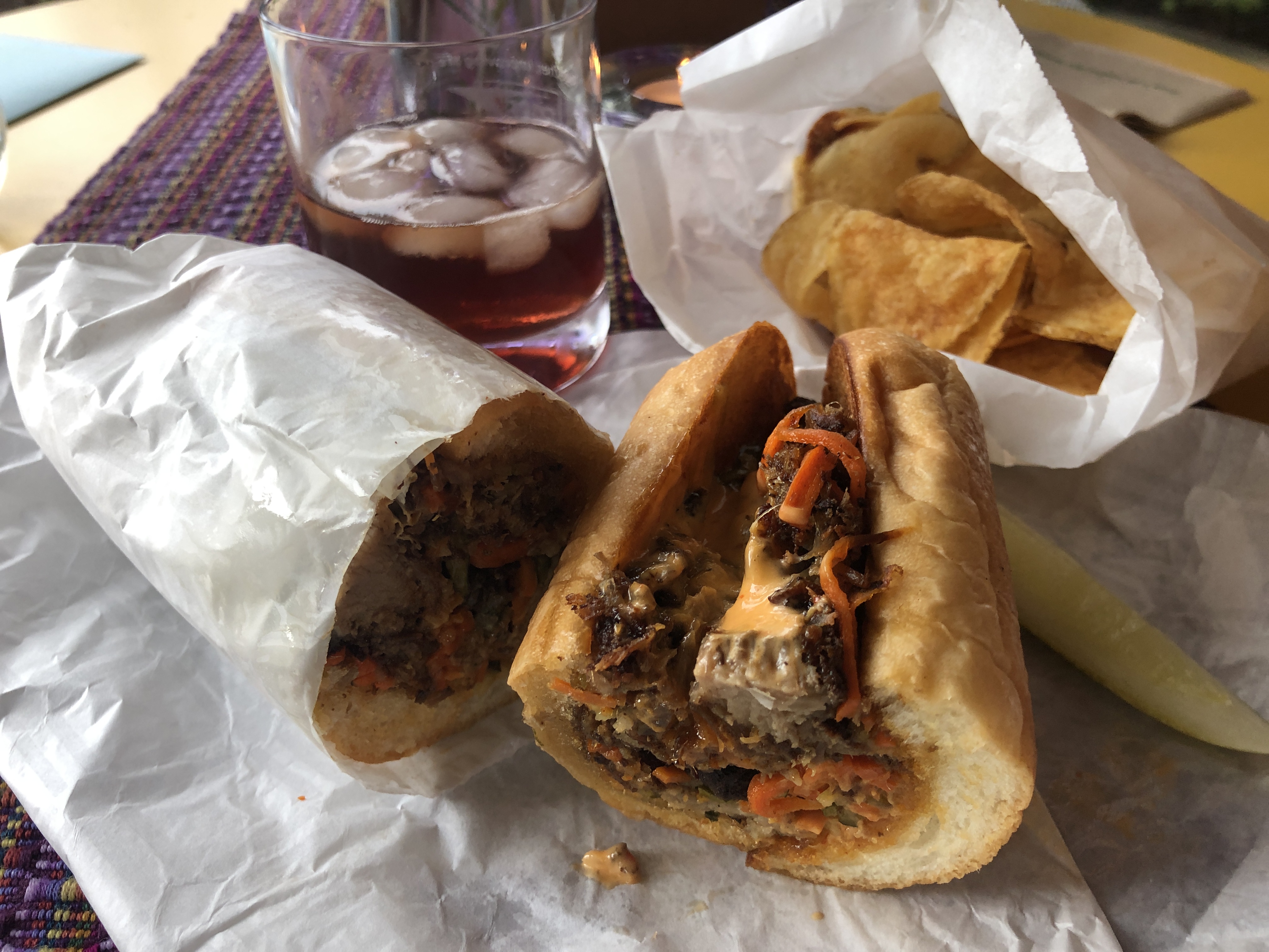 A soft hoagie roll holds a pile of browned meat layered with cheese and pickled onion and herbs. Beside it sit a white paper bag of homemade potato chips and a dark reddish brown cocktail on ice.