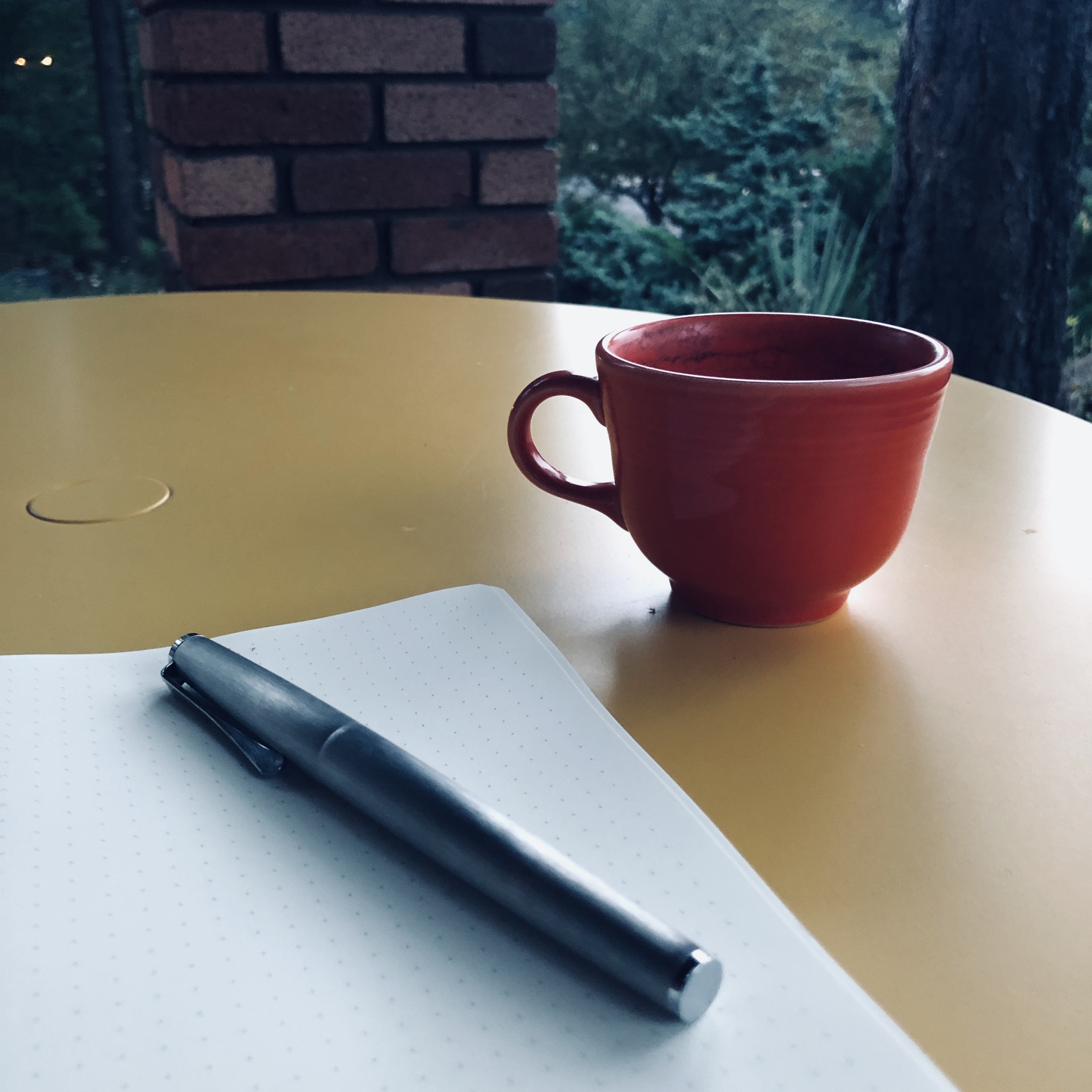 A small orange coffee cup sits in a yellow metal patio table, beside a notebook and silver pen. The dawn light is gray due to clouds. 