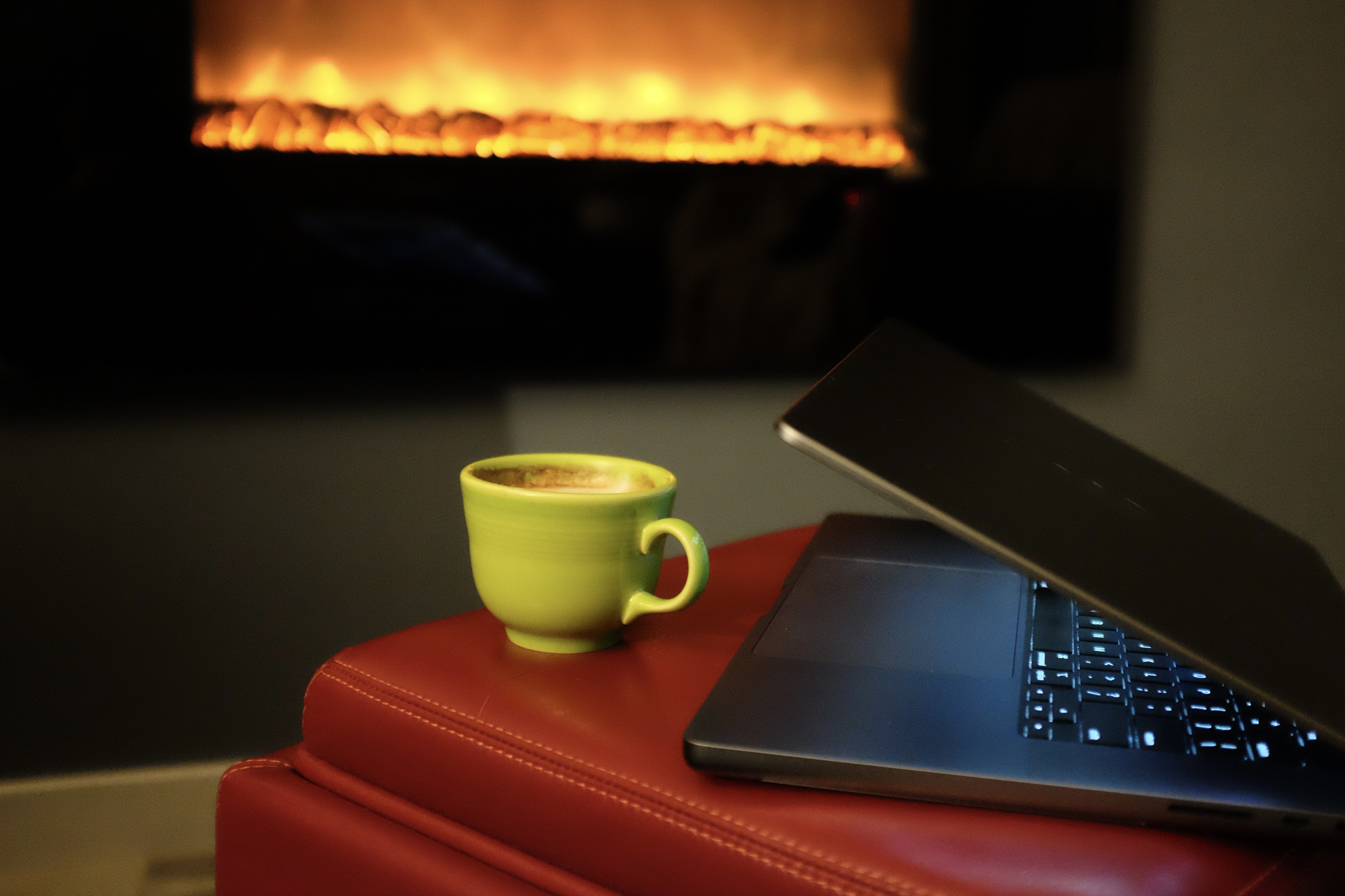 A close-cropped photo of a green coffee cup sits beside an open Macbook in front of a glowing, electric fireplace. 