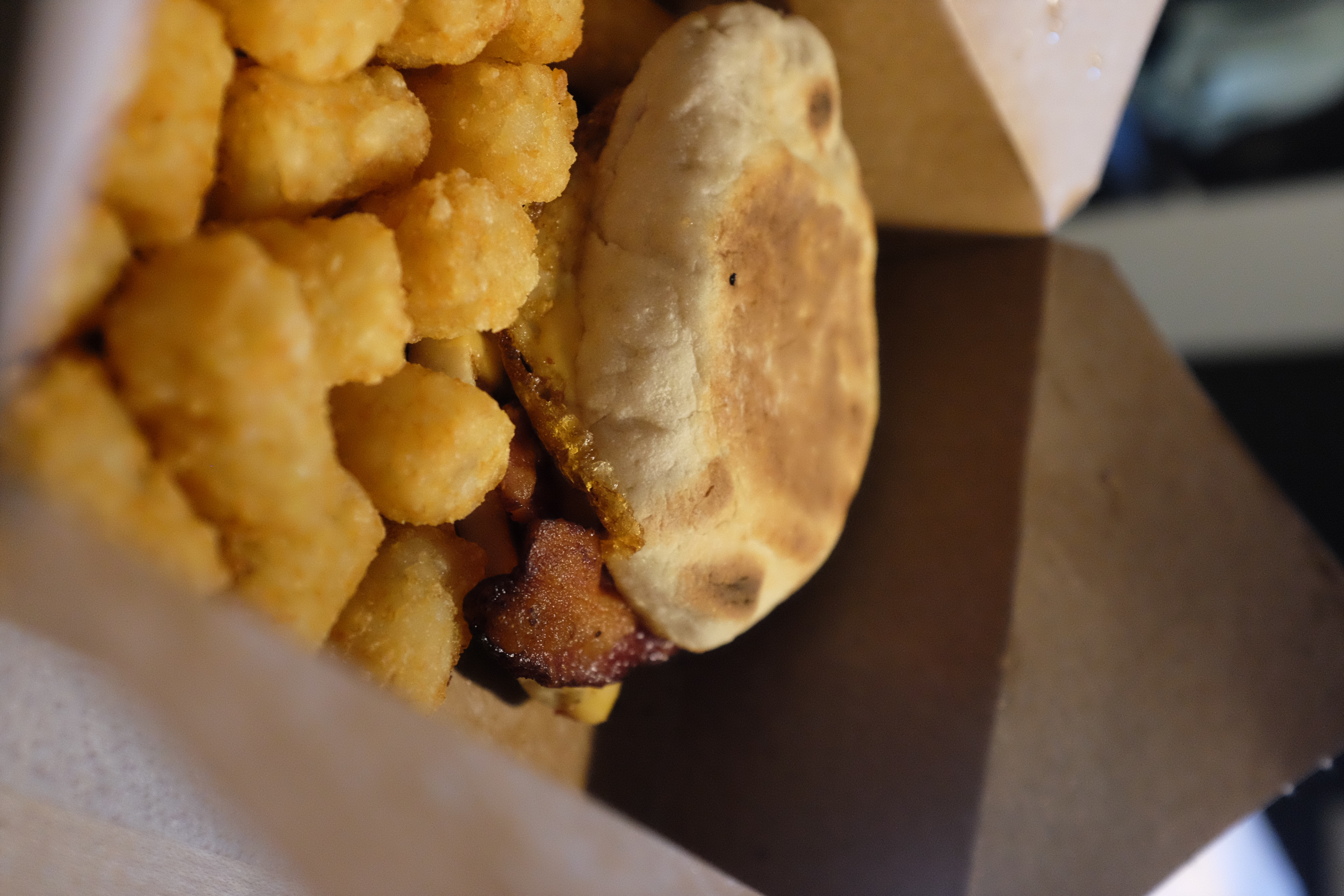 A sandwich of english muffin, egg and bacon sits in a take-out cardboard box, nested among a pile of tater tots.