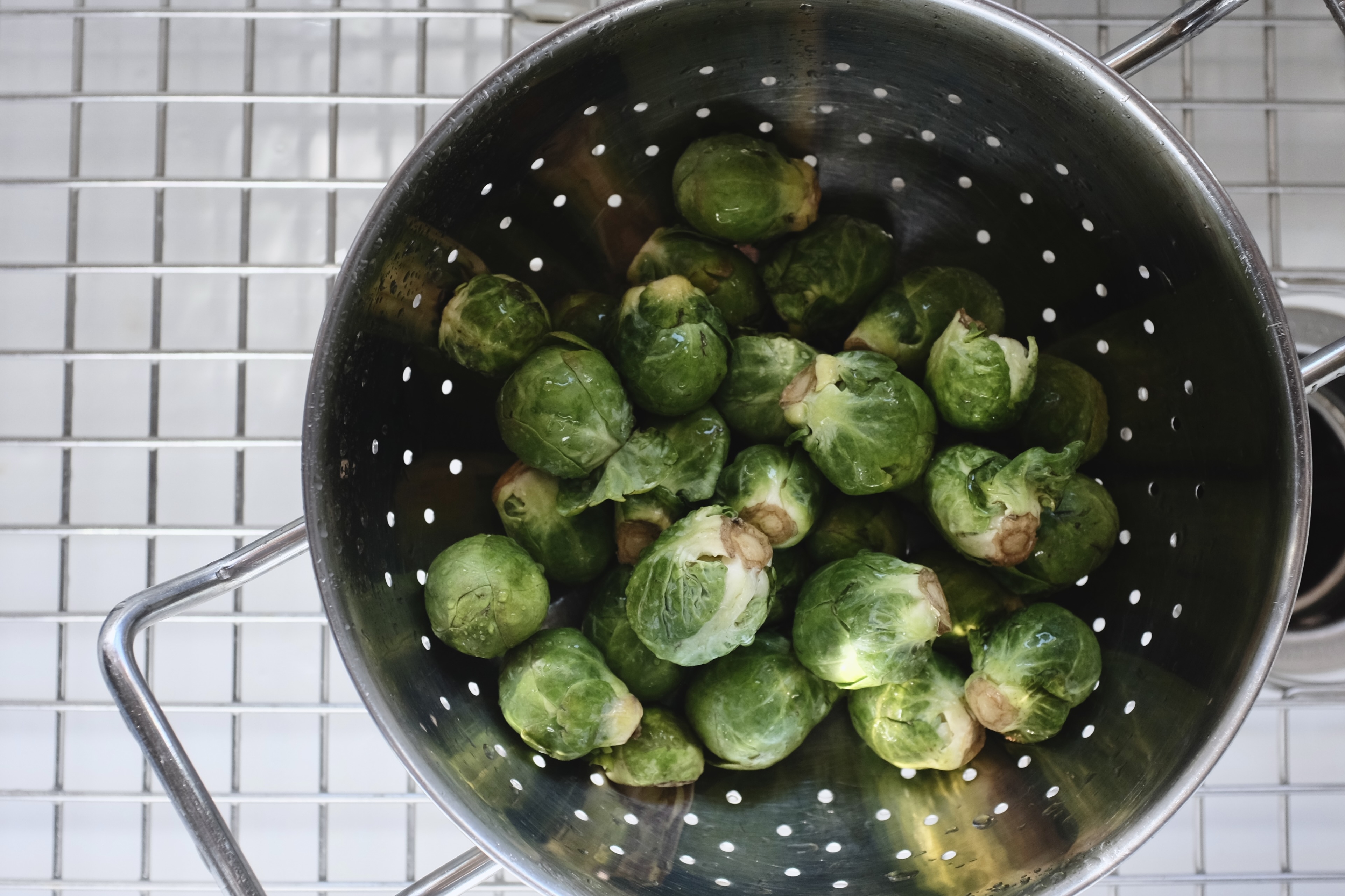 A pile of bright green brussels sprouts sit in a metal collander, in a white porcelain sink, photographed close and from above.