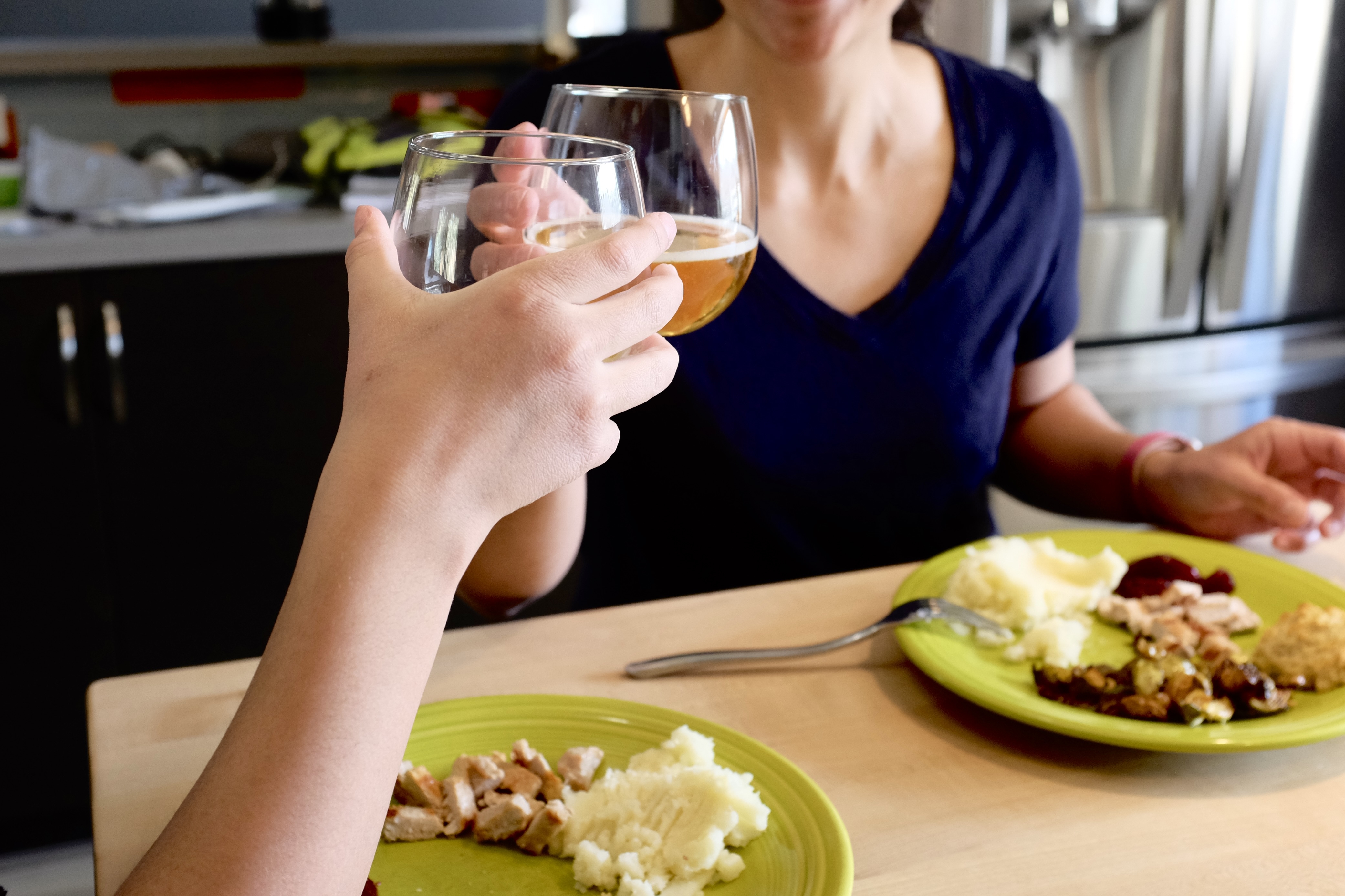 This photo is cropped so that two arms are seen touching a pair of glasses containing sparkling apple cider, over two green colored plates, holding slices of pork, mashed potatoes, and brussels sprouts.