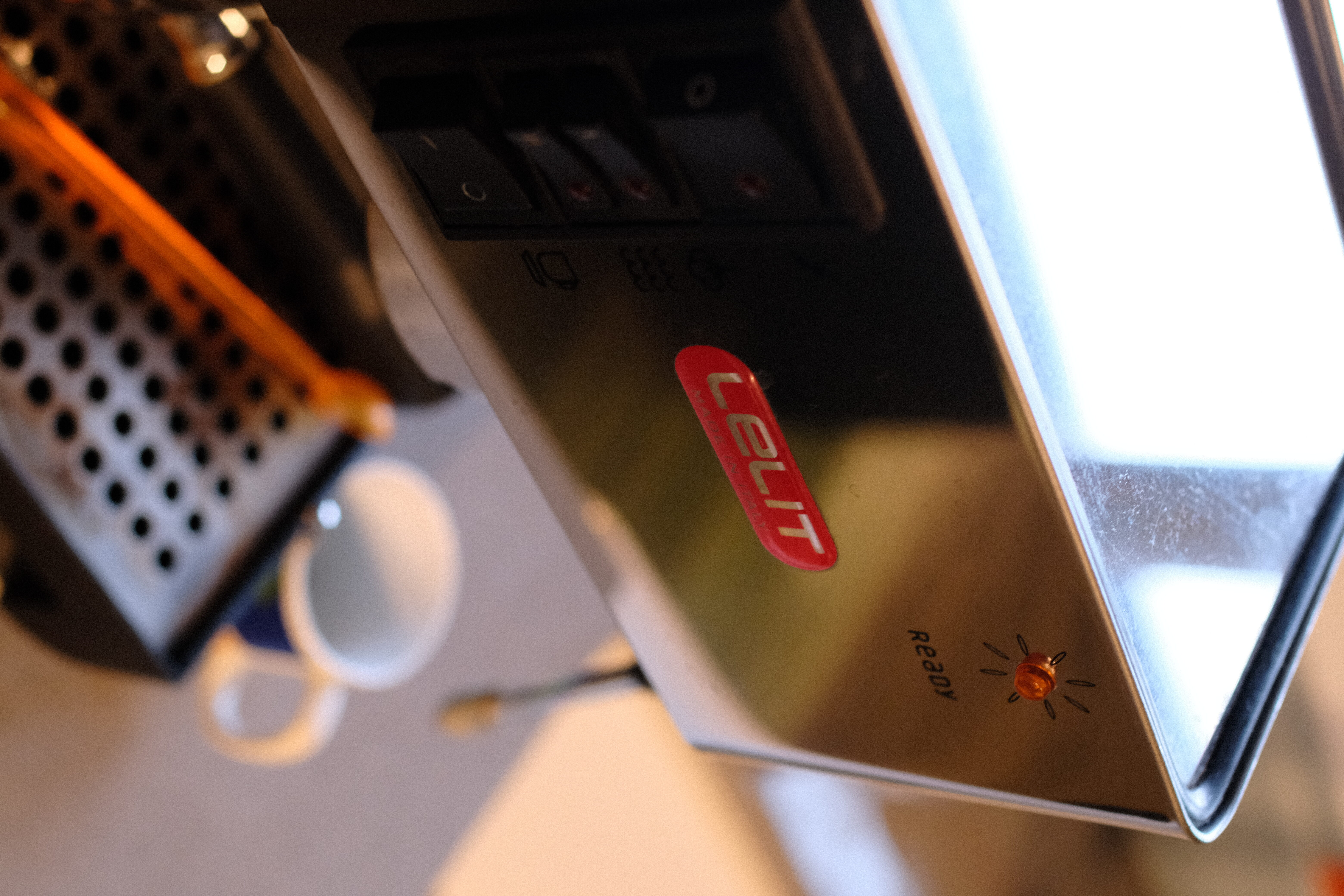 A Lelit espresso machine photographed at an angle and showing a narrow depth of field with a blurred, small cup beside it.