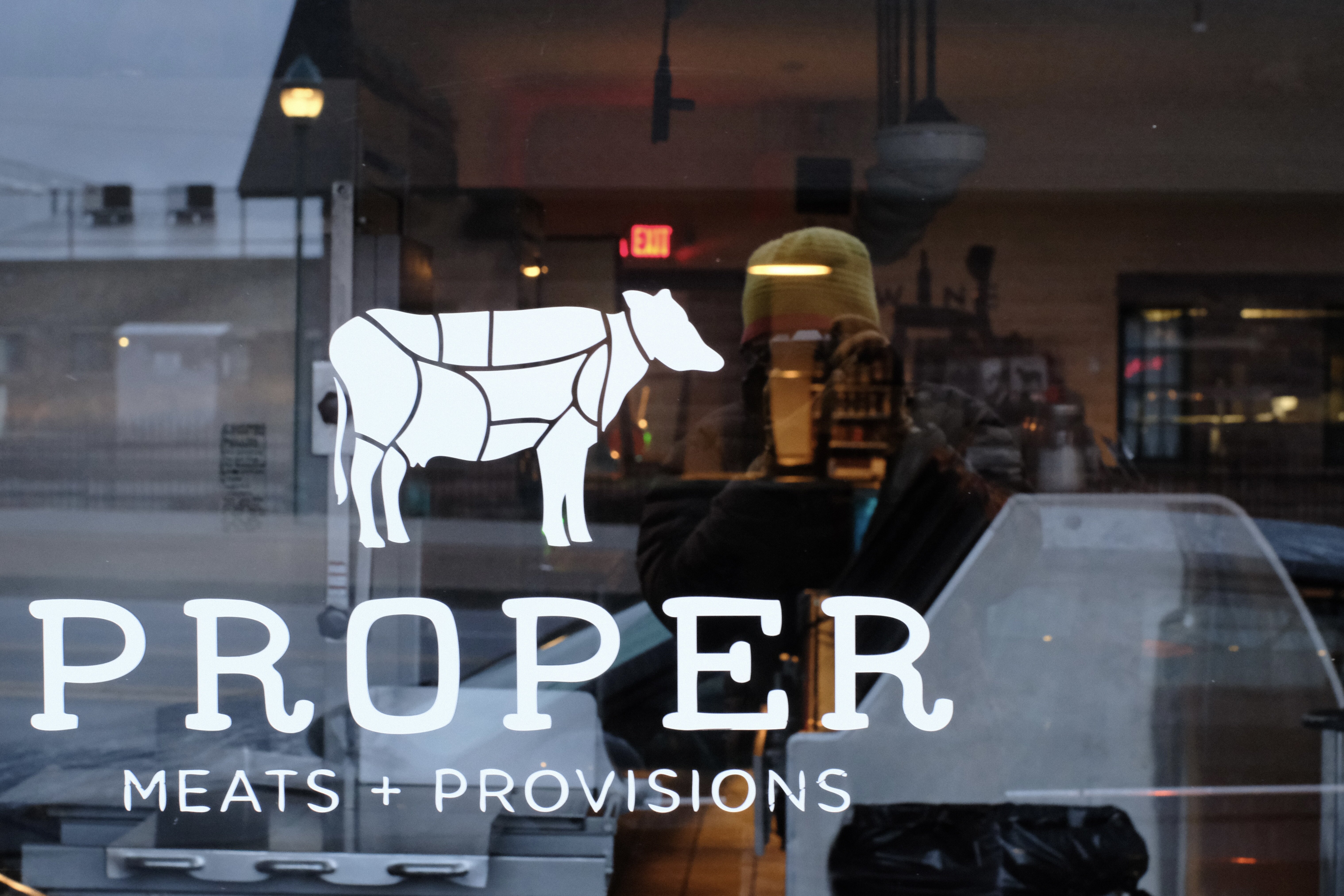 A large plate glass window with stylized printing reading ‘Proper Meats and Provisions.’ A photographer in a yellow hat, holding camera to his face, is visible in the reflection.