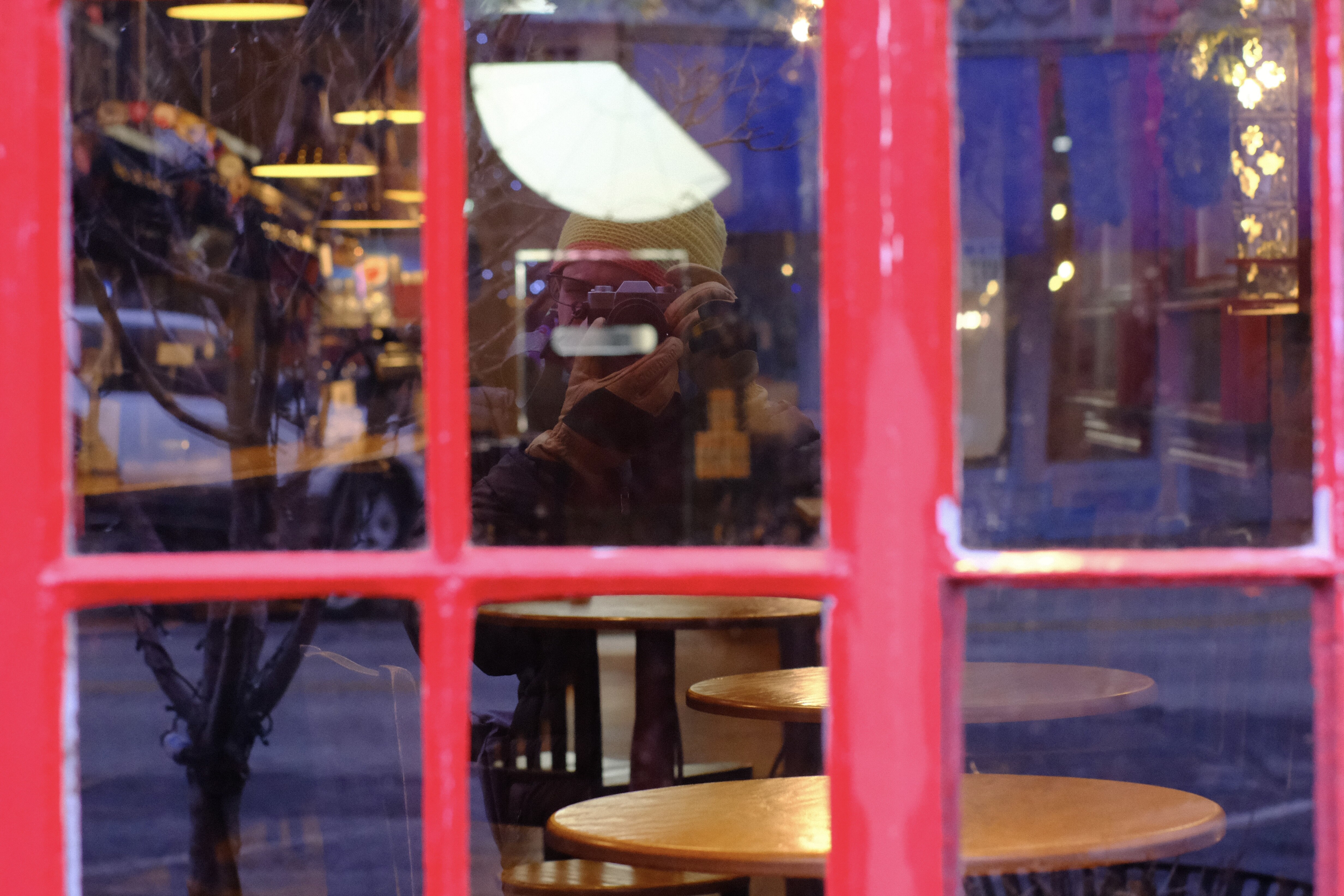 A set of bright red windowpanes frame a reflection in the glass of a photographer with a silver camera to hit face. He is wearing a yellow knit hat and yellow leather gloves. Behind the glass several chairs and a long bar counter are visible in warm indoor light.
