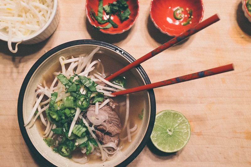 A bowl of soup sits on a wooden counter. The broth is brown and has a pile of steak, scallions, bright green jalepenos and bean sprouts on top. A pair of bright red chopsticks sits in the bowl. Several small red dishes of garnishes sit above the bowl and a sliced lime is beside it.
