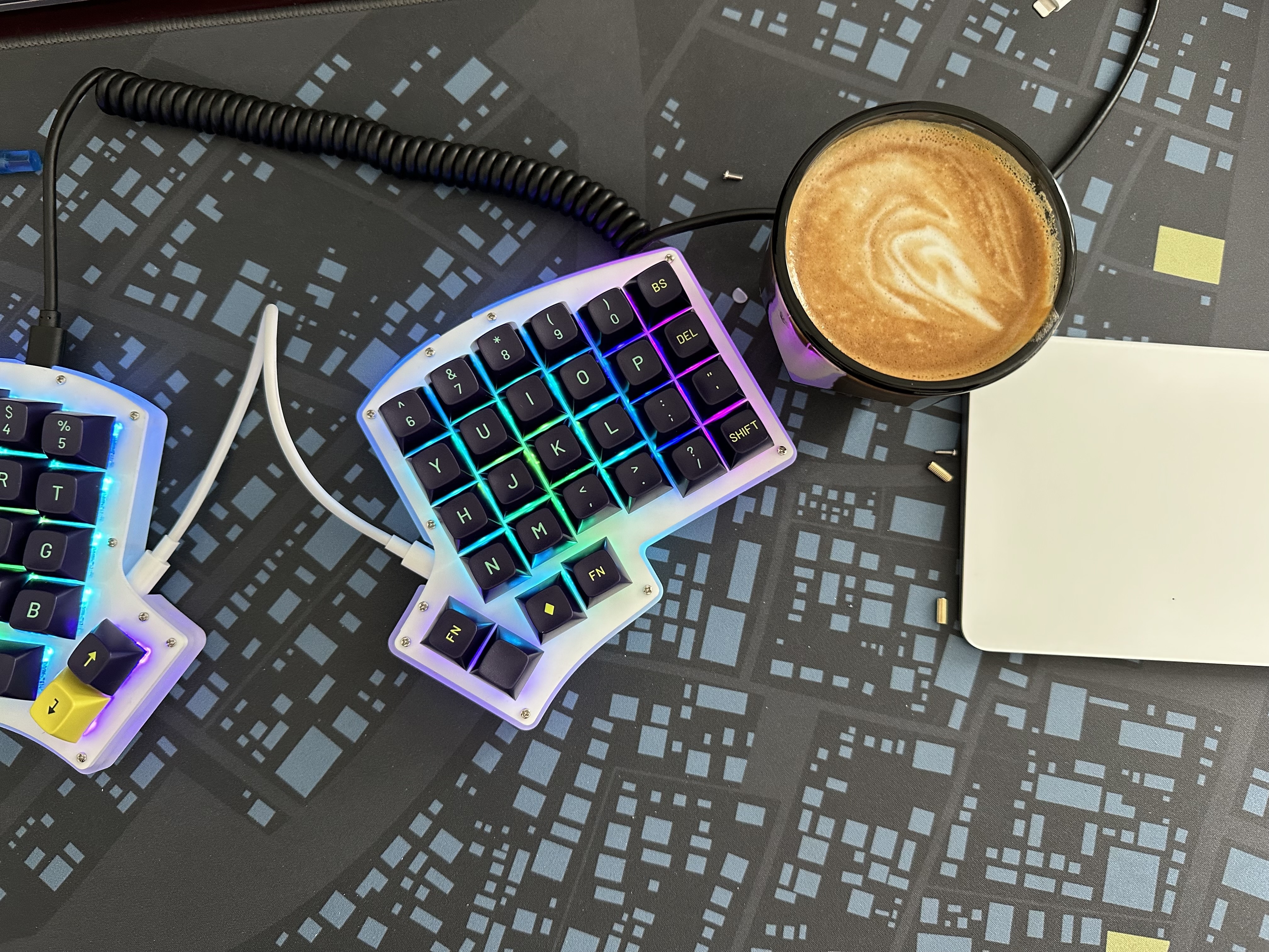 A top down view of a split ergonomic keyboard, the Keeb.io Iris. It has backlit keys on a white plastic top plate and dark blue keys with cyan legends. Beside the keyboard is a mug of coffee with a swirl of frothed milk on top, and an Apple Magic Trackpad sits next to the coffee.