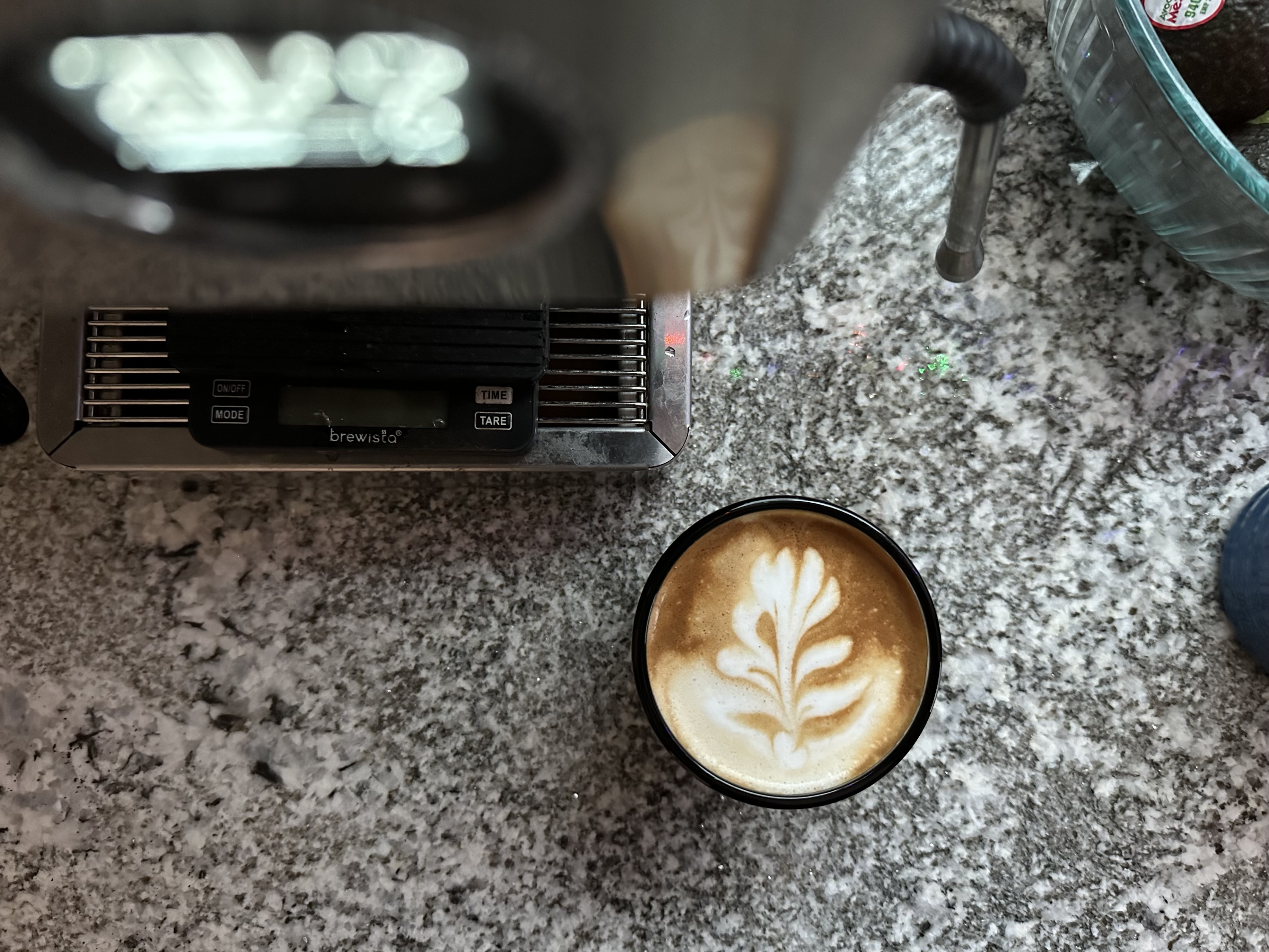 A top-down view of a cappuccino in a smokey-gray colored glass cup. It sits beside the drip tray of an espresso machine which, and out of focus in the top of the frame is the small digital display of the machine. The cup and machine sit on a speckled black and white marble counter and natural light comes across the frame from the top. 