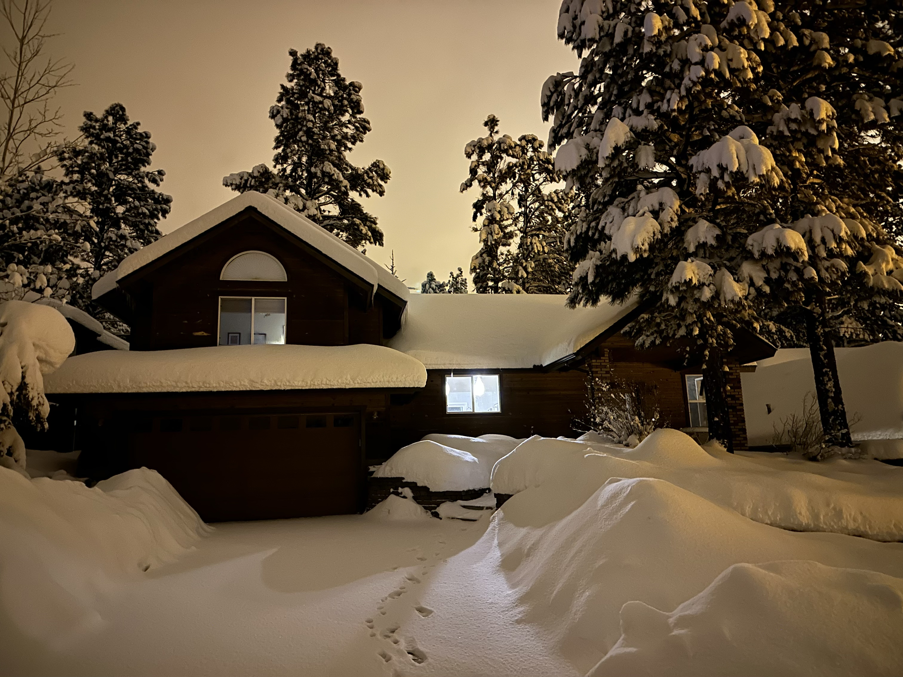 A brown wood-sided house seen at night. Its roofs and areas next to the sidewalk and driveway are piled with high snow. Lights shine in the front windows and the cloudy sky is lit palely by ambient city lght. 