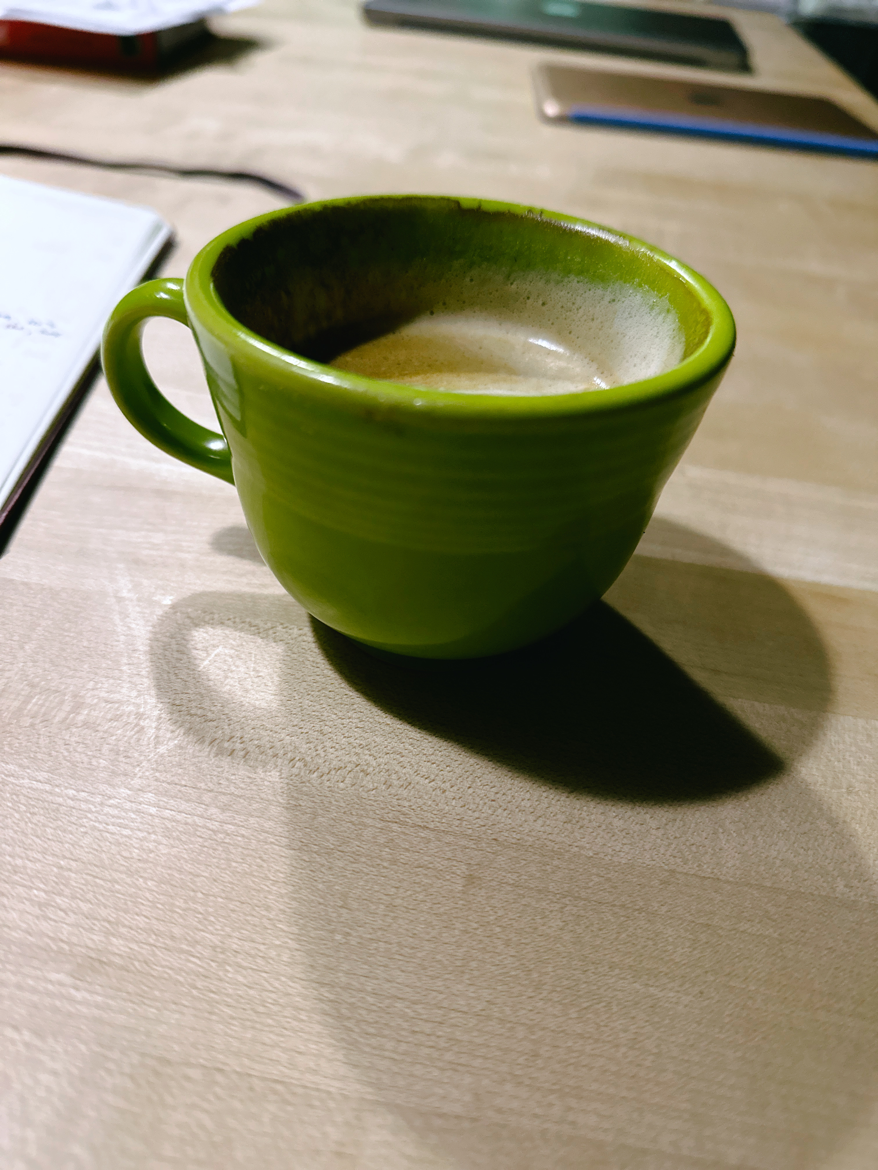 A green, half full cup of milky coffee, in a wooden countertop beside an open notebook. 