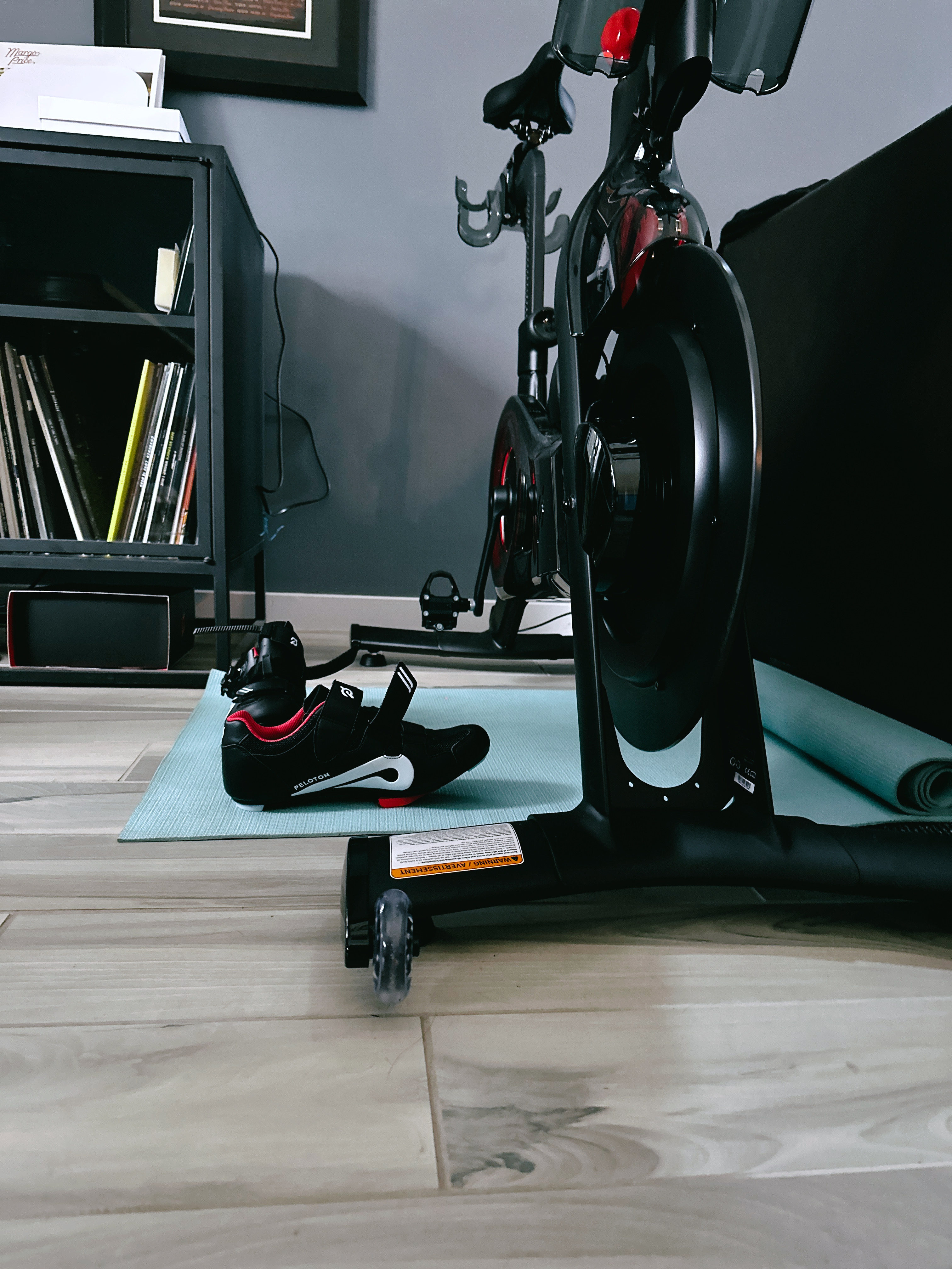An indoor bicycle, a Peloton, photographed from the front at a low angle. A pair of black cycling shoes sit next to it on a light blue mat. 