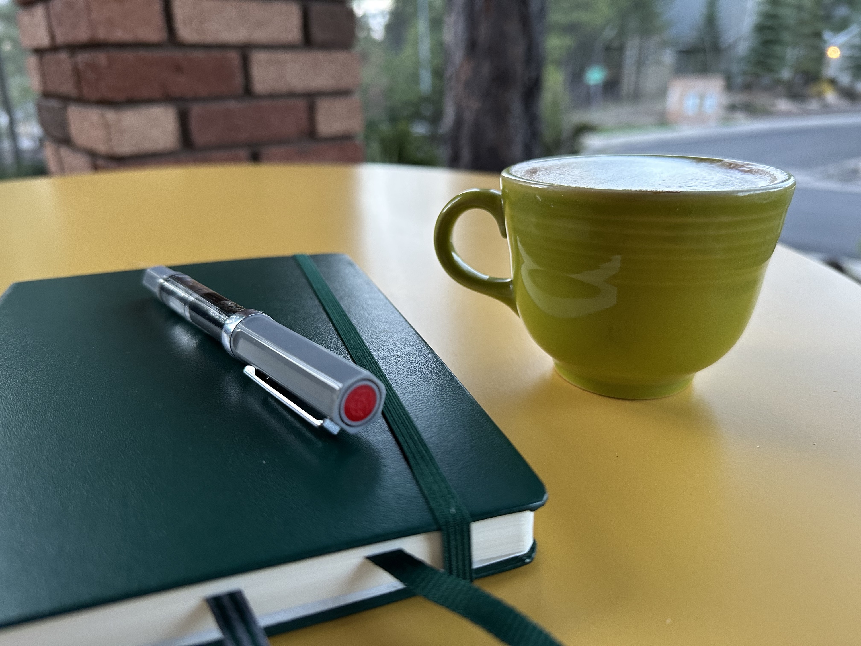 A pen, leuchtturm notebook and green coffee cup sit in a yellow outdoor patio table. 