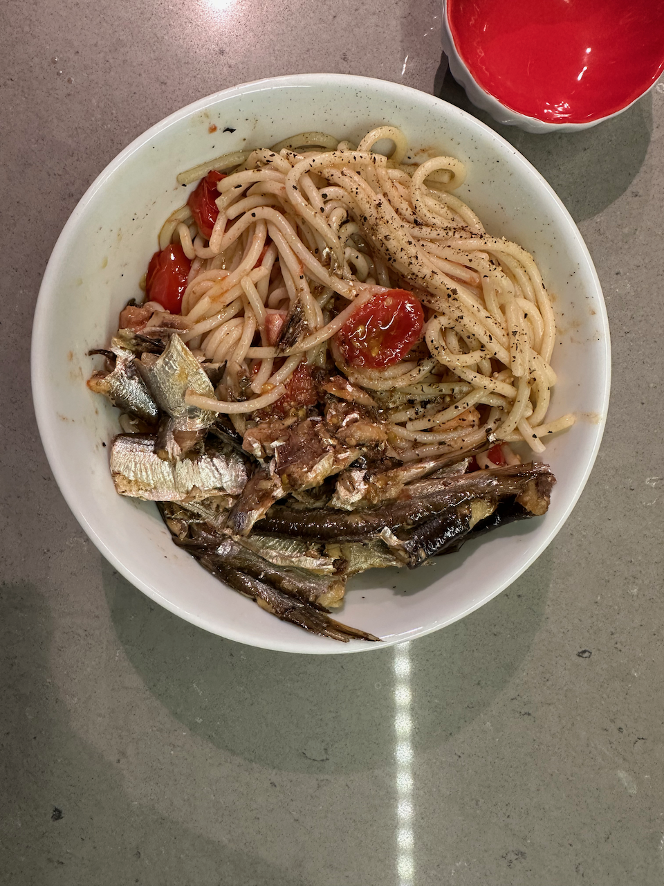 An overhead photo of a white bowl on a gray countertop. Spaghetti noodles fill most of the bowl and are topped with lightly cooked, slices of cherry red tomato. Beside them are several slices of small, silvery sardine fillets.