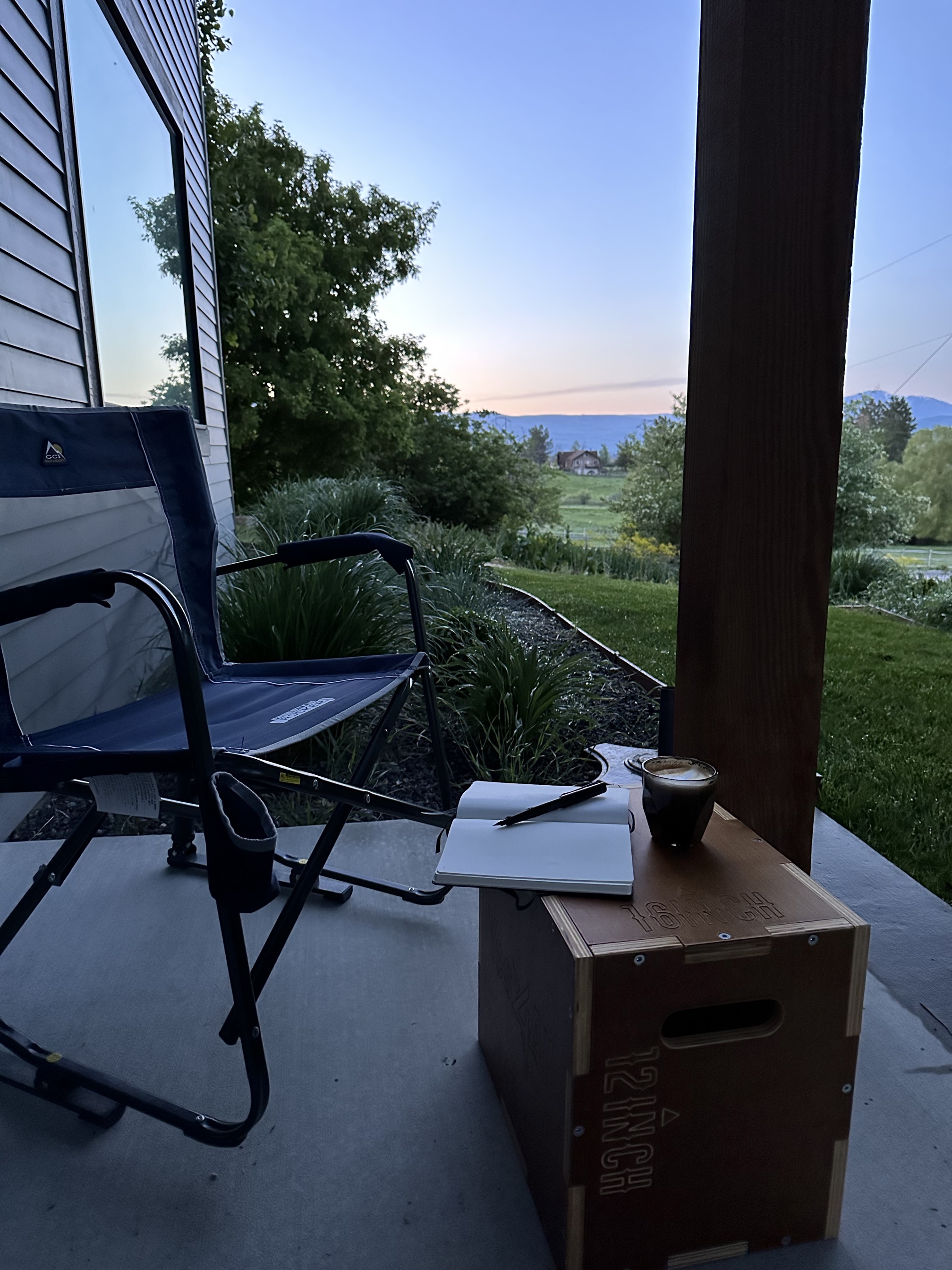 A wooden box sits on a small porch. On top of it are a coffee cup and an open notebook with a pen. The sunrise is showing just a bit of color far in the background, reflected in a large window beside the box. 