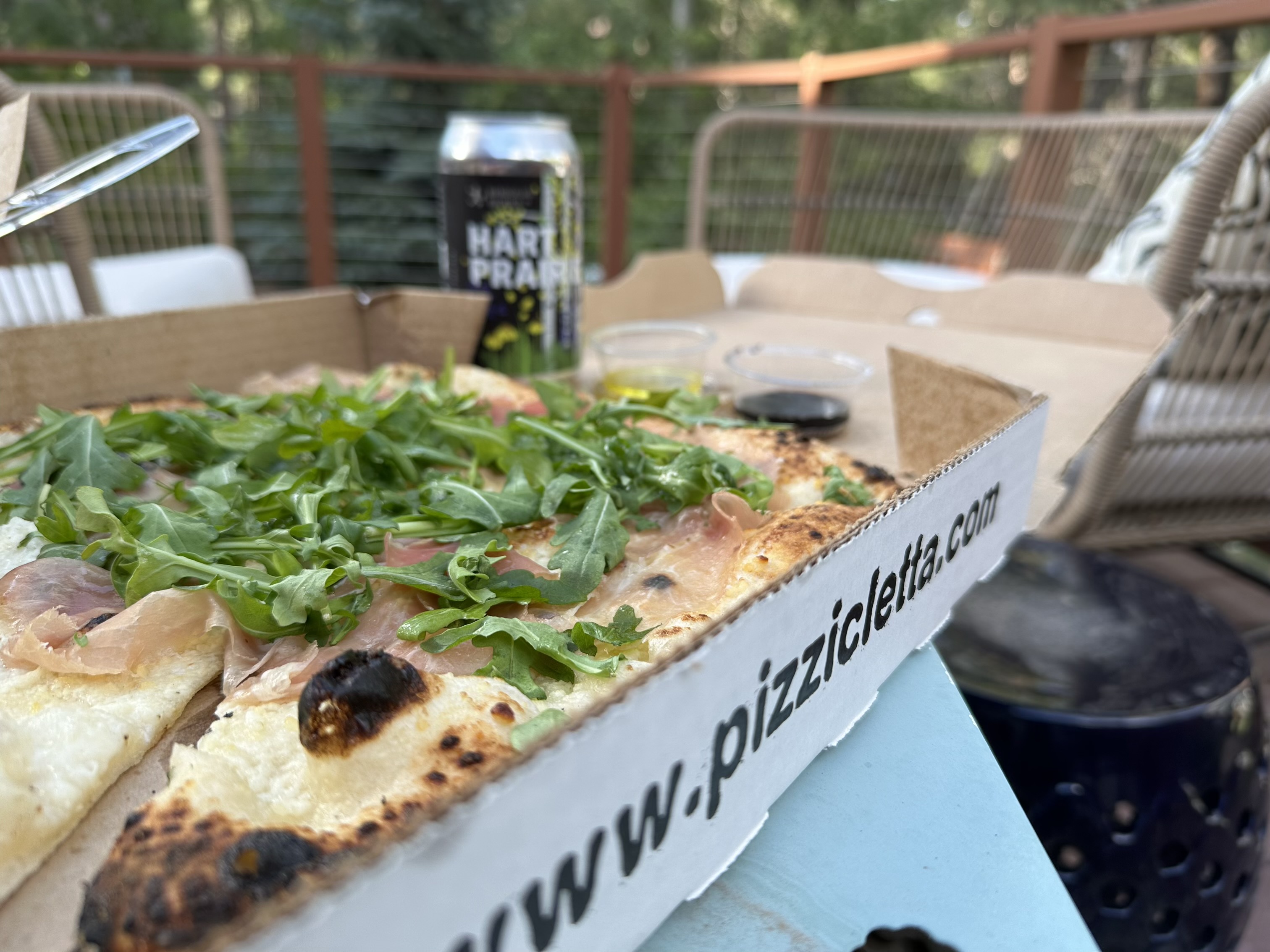 A pizza box sits on a metal outdoor table. The pizza has arugula and airy slices of prosciutto, and charred puffs of crust. The box reads ‘pizzicletta’. Beyond the box is a beer can. 