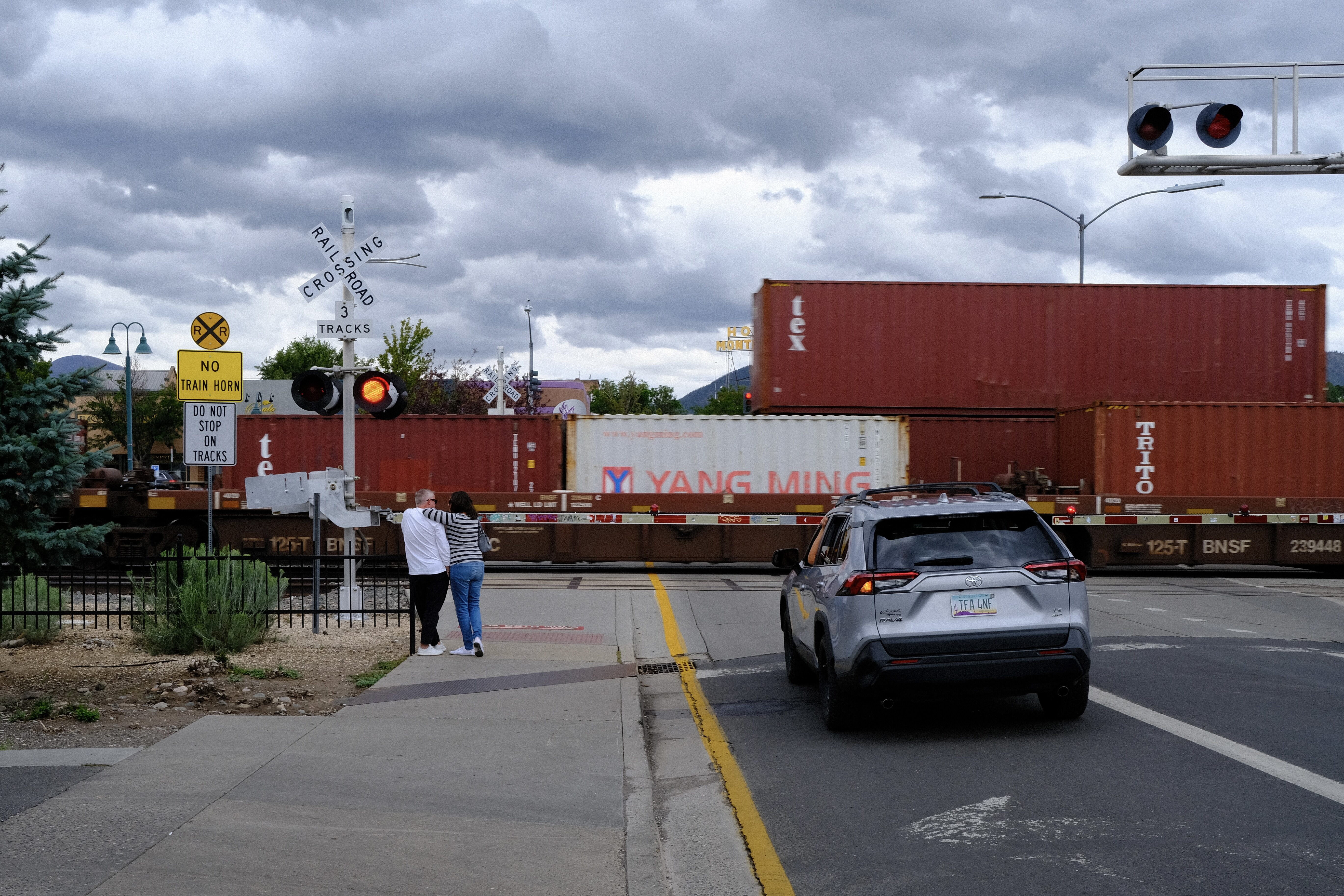 A person leans lightly against a railroad crossing signpost, facing away. Another person leans slightly on them. The crossing gate is down and two trains are passing, in opposite directions, on the tracks ahead of them.