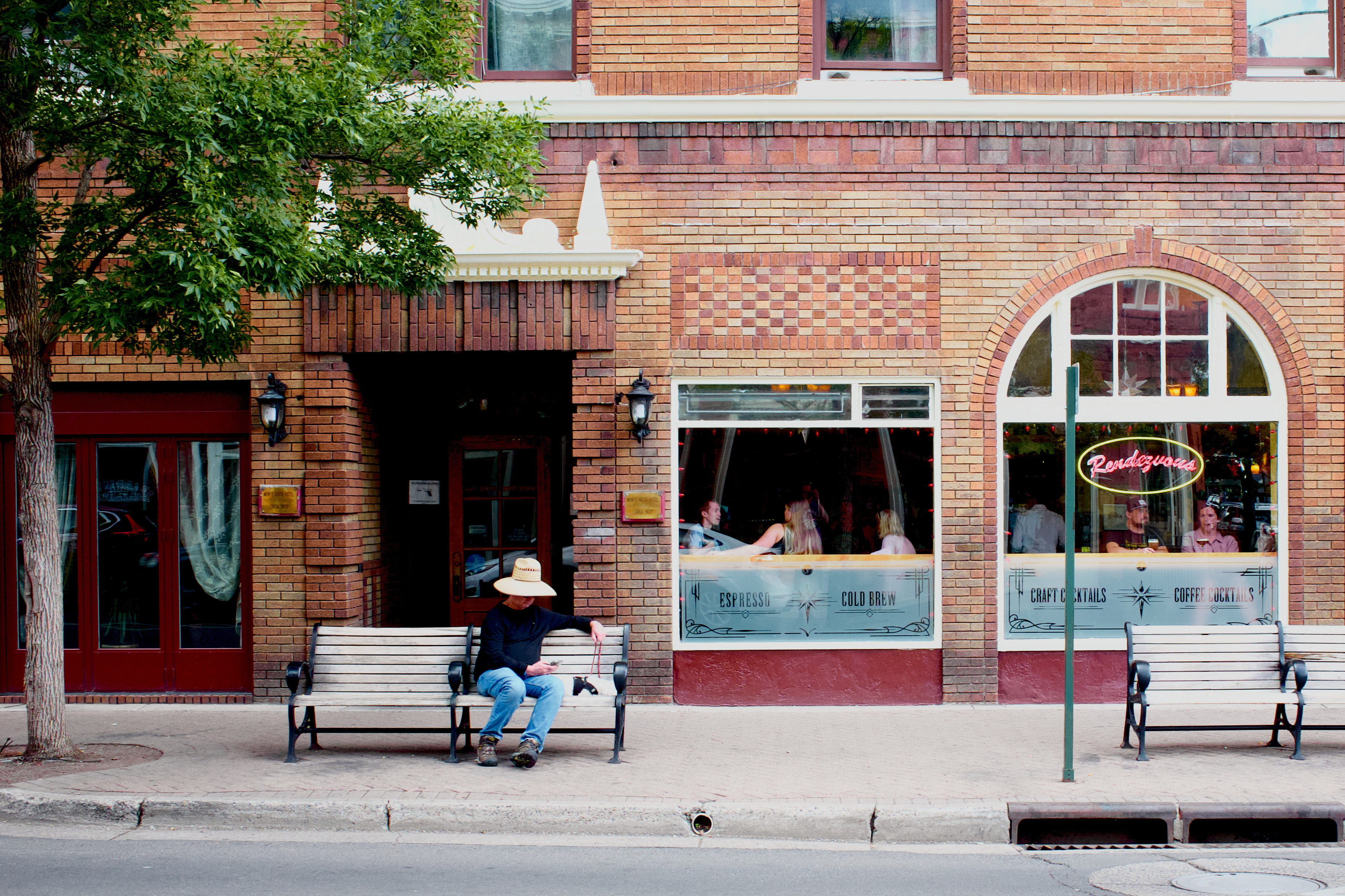 A person sits on a worn wooden bench outside a brick building, across the street from the photographer. Through the windows several people can be seen gathered, talking. There is a red neon sign in one window reading ‘Rendezvous’ in a cursive font. 