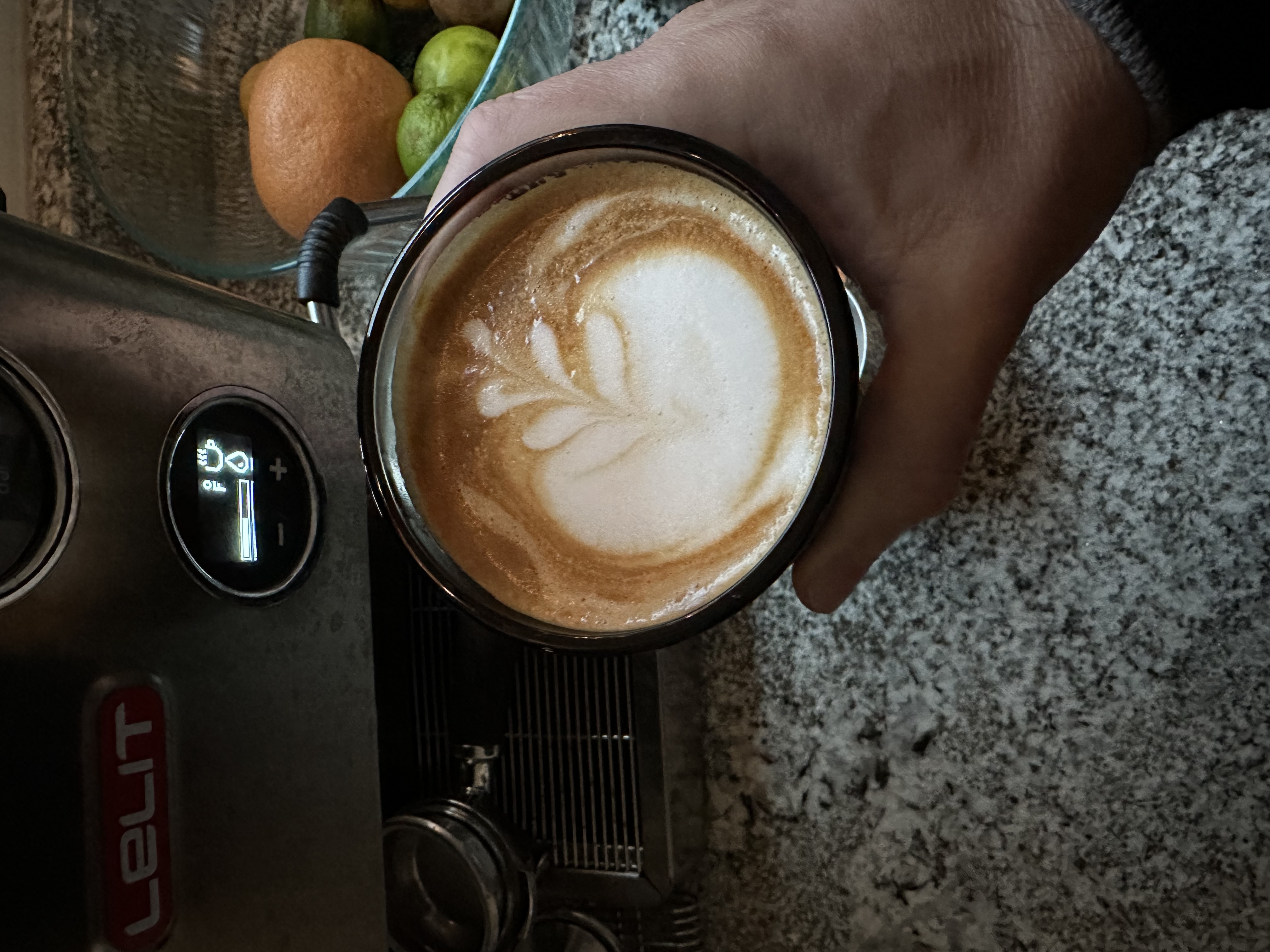 A hand holds a gray glass of cappuccino above a speckled black and white countertop. The cappuccino is a brown sugar color with a bulbous and somewhat accidental looking floret of steamed milk poured on top.