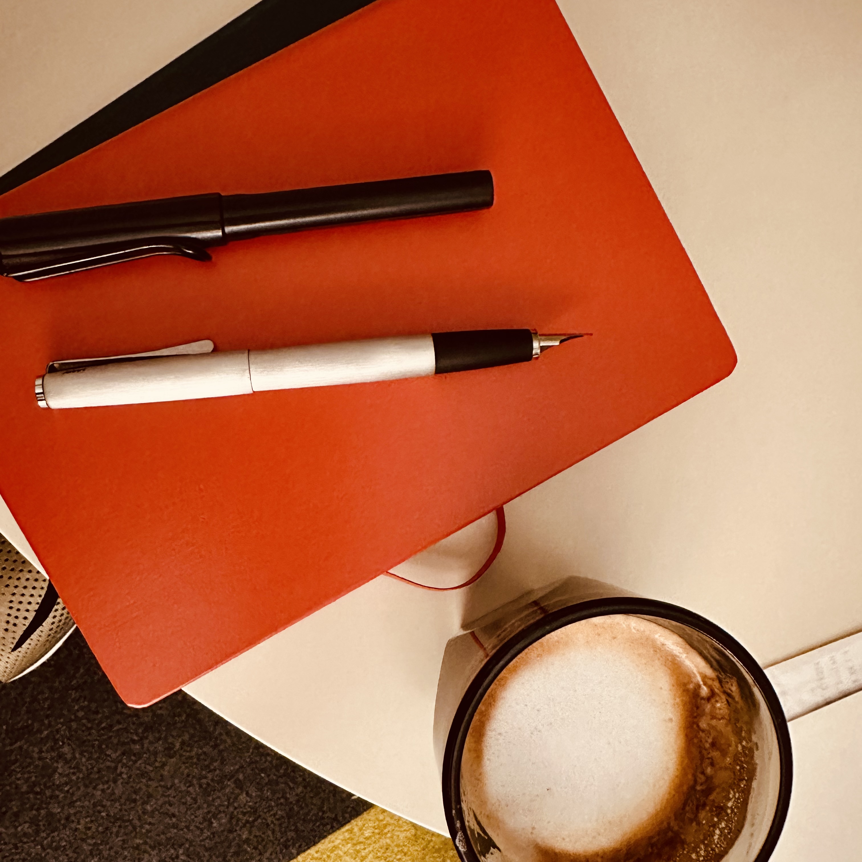 Two fountain pens sit on an orange hardbound journal. One pen is silver colored, a Pilot Studio with black barrel with its cap posted. The other is a black Lamy Safari. Beside the journal is a half full cup of cappuccino. 