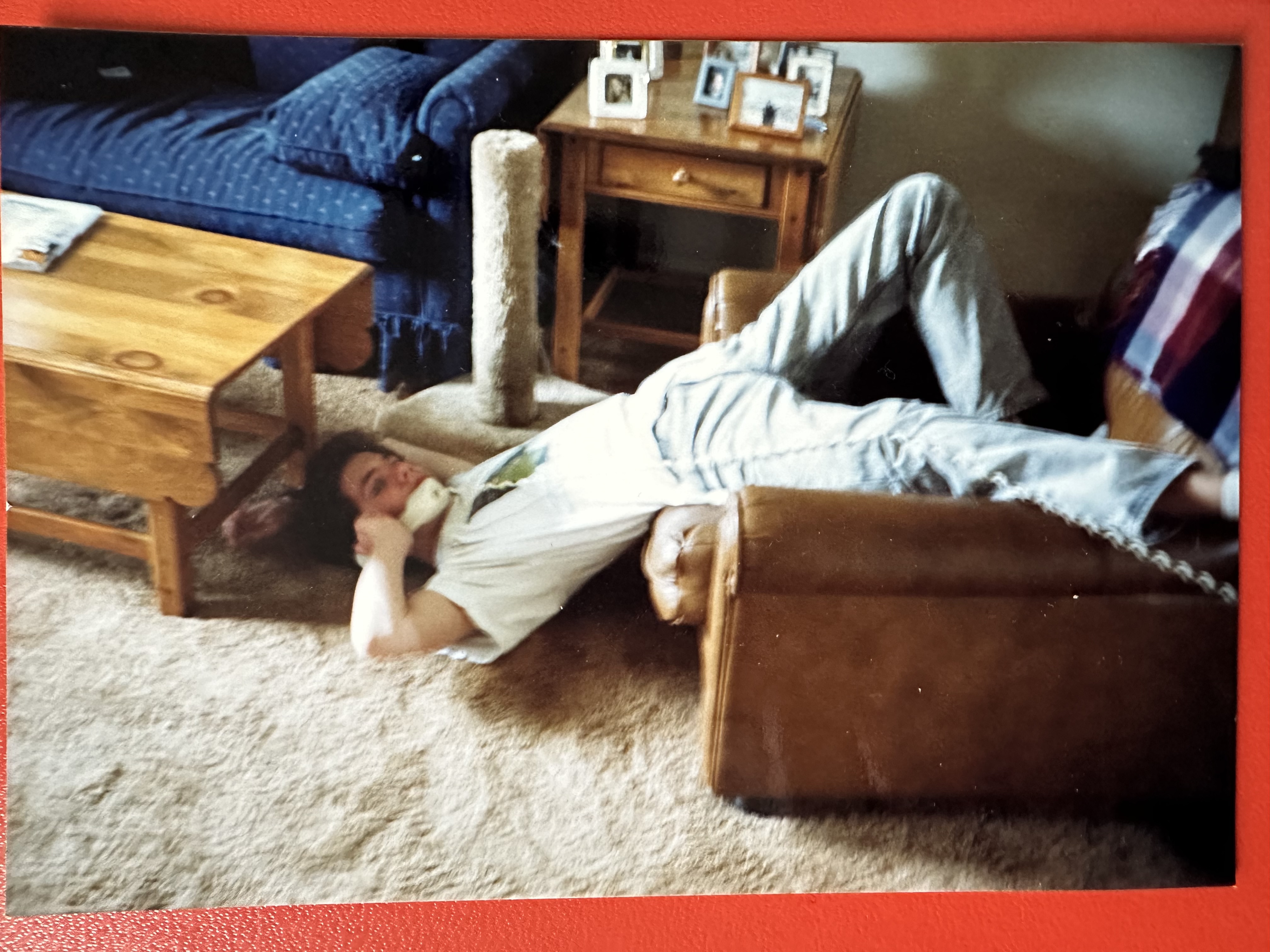 A teenager lays upside down in an armchair, his head and shoulders on the carpet. He is holding a phone receiver to his ear and the cord stretches along his body and out of frame. 