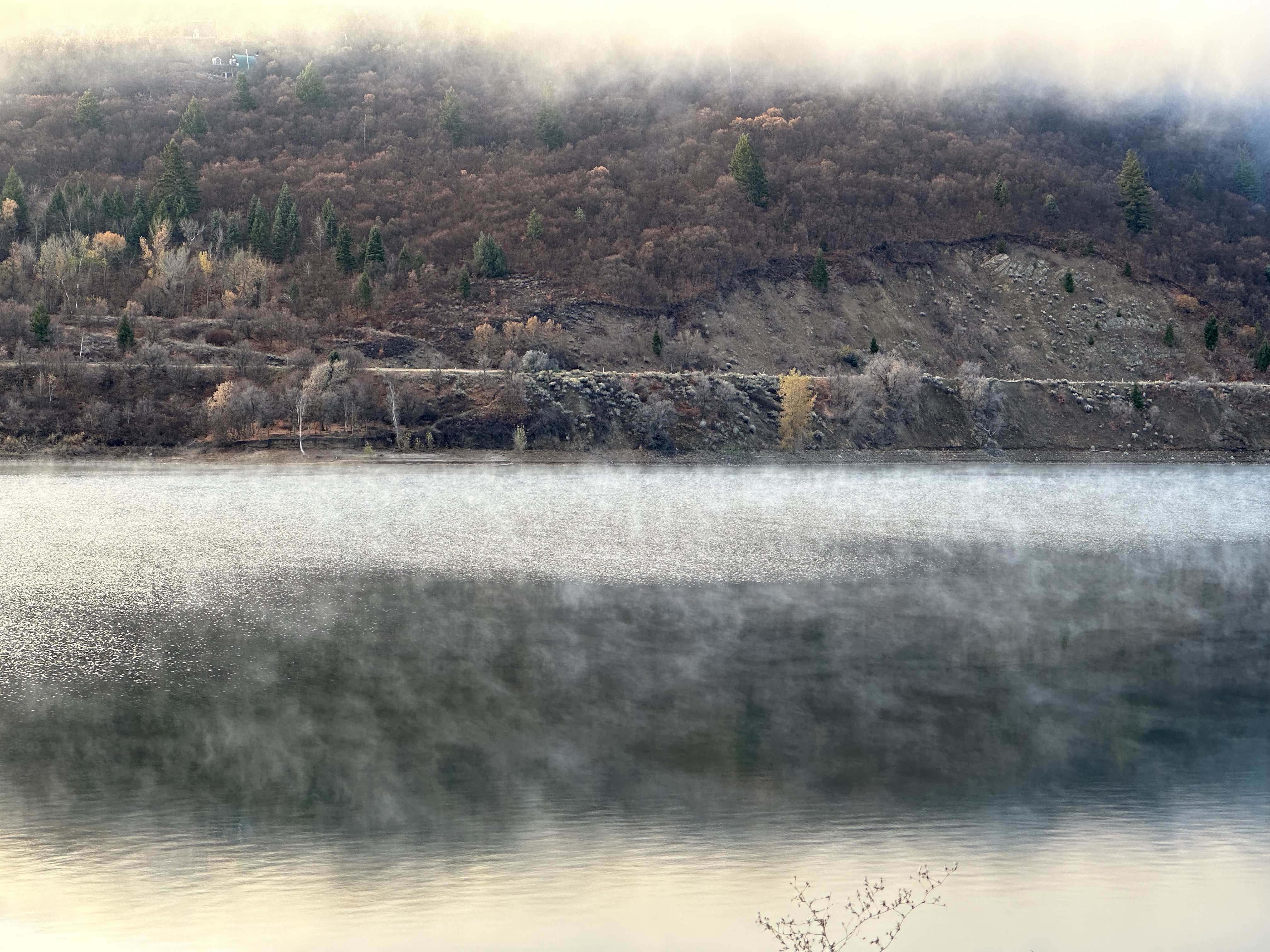 A photo in grey and subdued autumn brown, showing a reservoir against a hillside, with low clouds at the top of the frame reflected in water at the bottom of the frame.