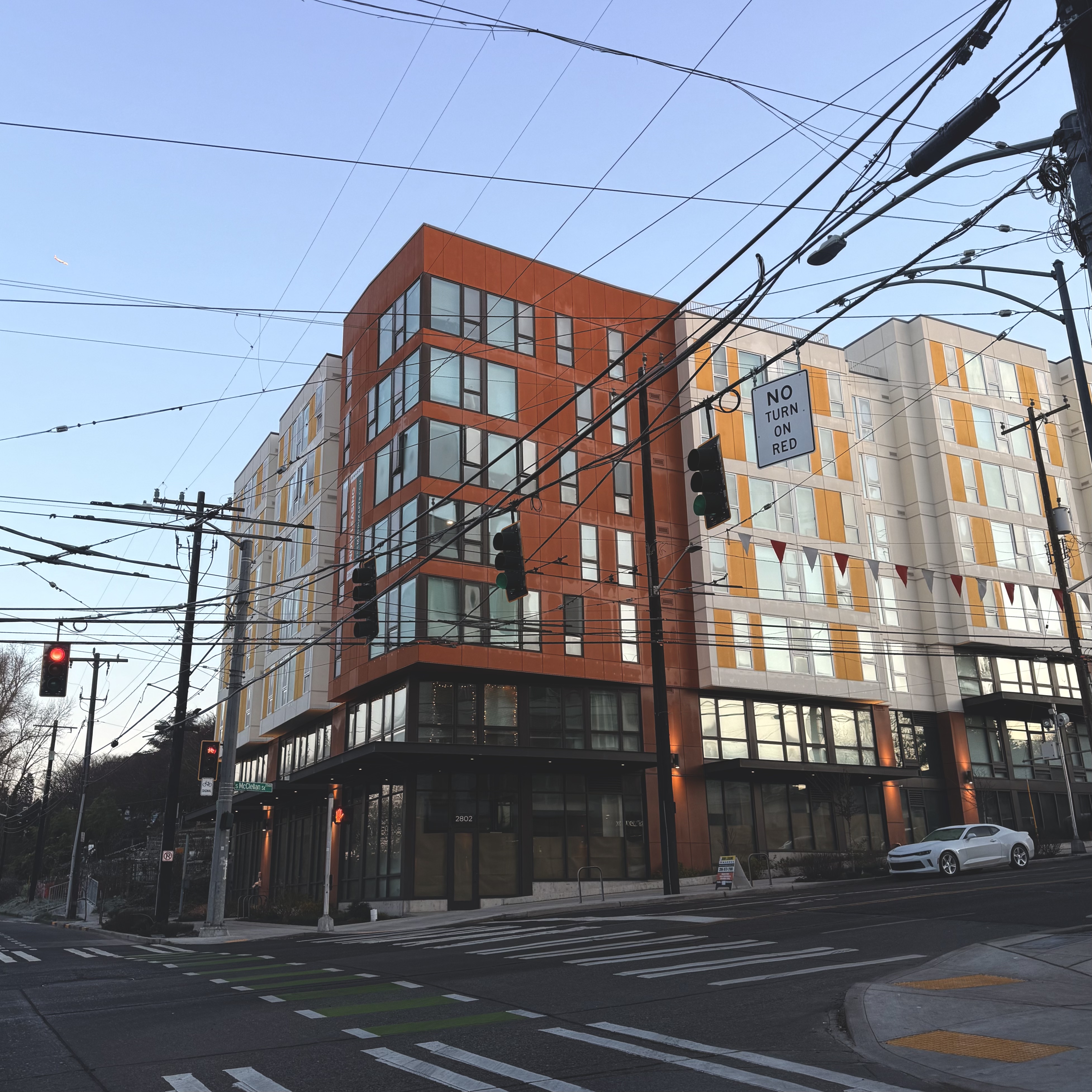 An intersection flanked by six-story apartment buildings in red, white and yellow paneling. Their windows are opaque but bright with morning, low-angle light. The intersection is criss-crossed by utility wires and cables for electric buses.