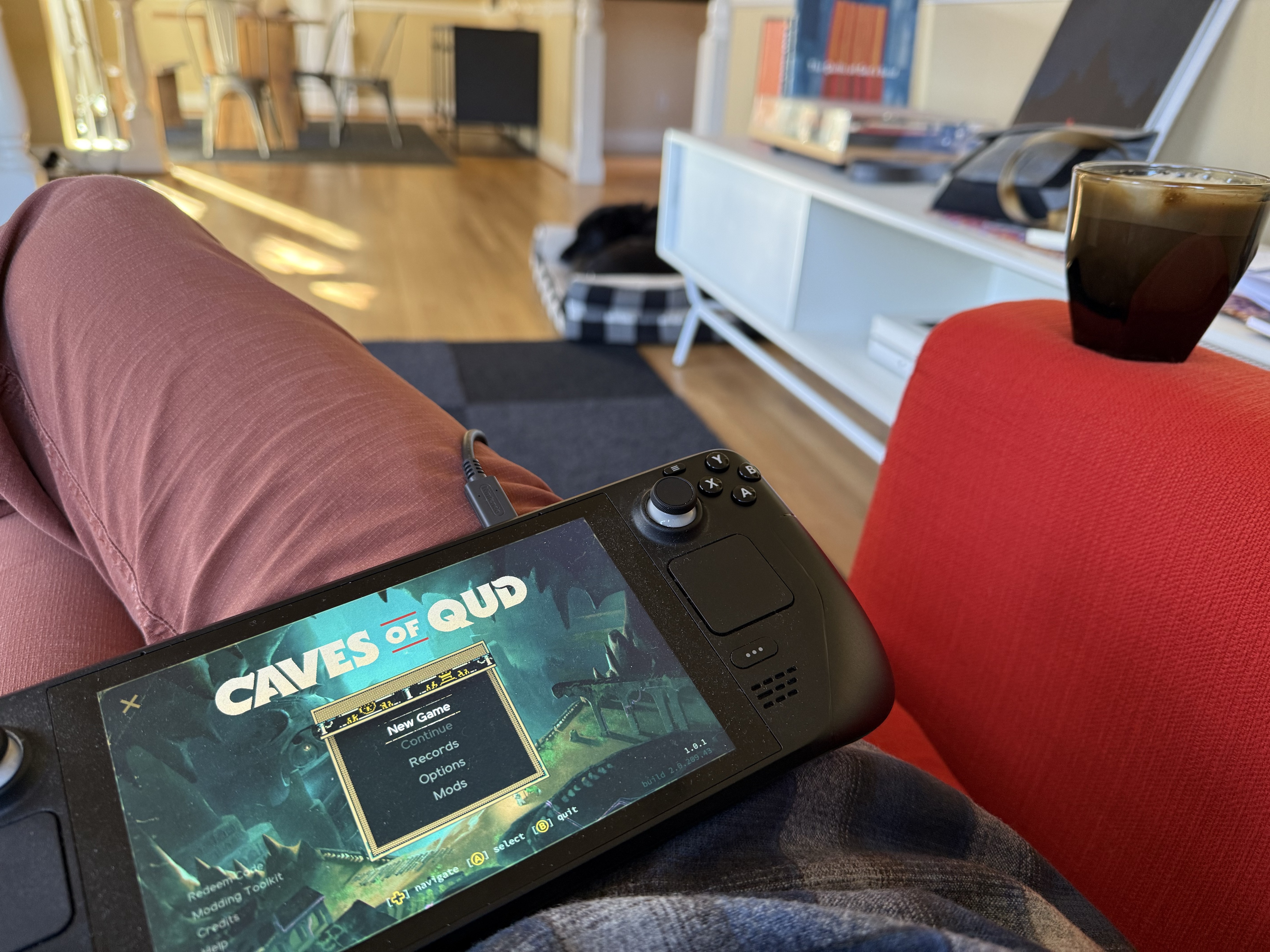 Light shines on a hardwood floor in my living room. The photo shows my legs in light red colored pants with my Steam Deck on my lap. The Deck is showing the Caves of Qud opening screen. On the arm of the bright red-orange chair is a cup of coffee. Out of focus in the background, my black dog lies in her bed. 