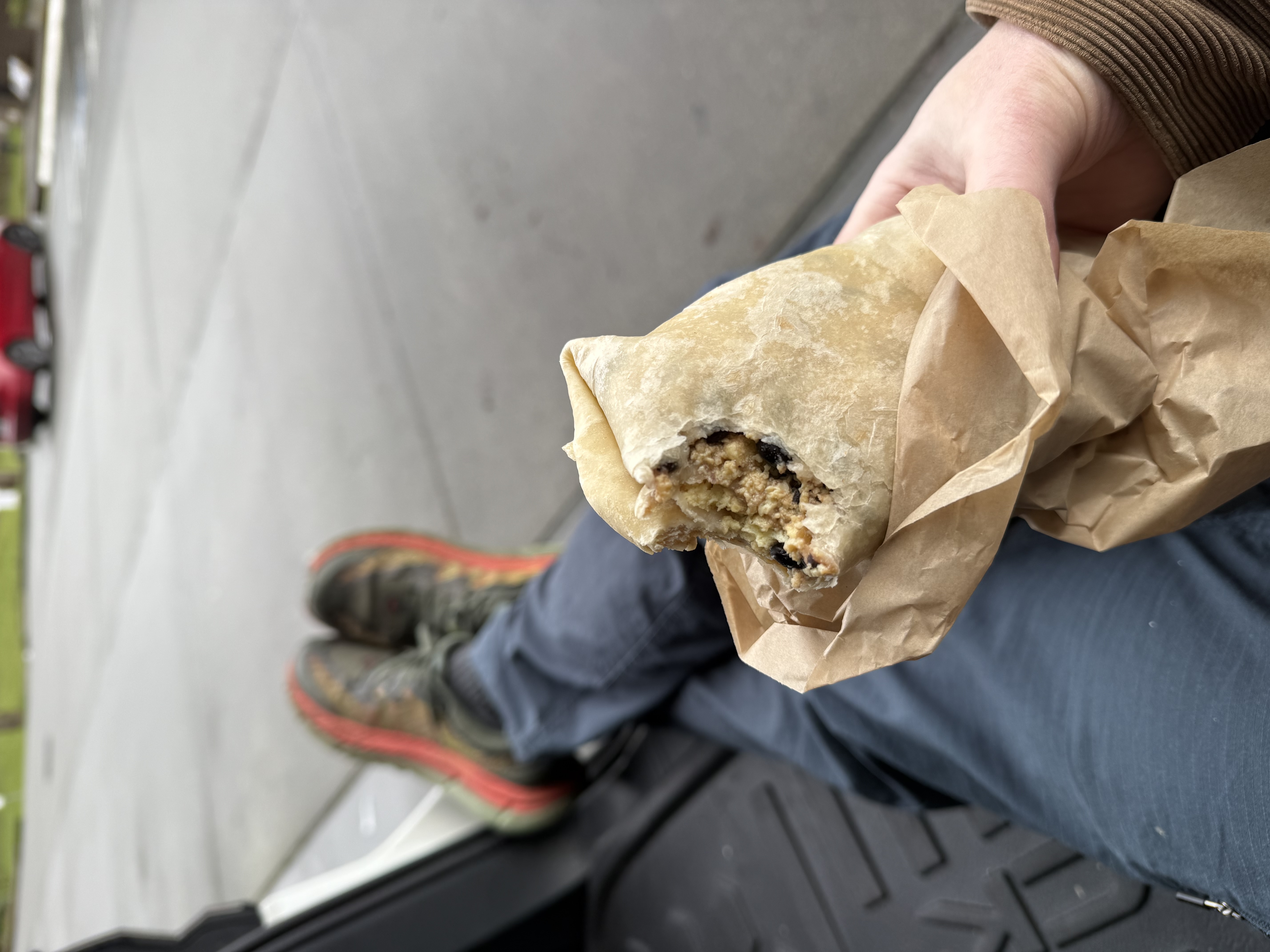 My hand holding a breakfast burrito, the paper partially unwrapped to show the tortilla and some egg and bean filling. I’ve taken a bite. In the background are my legs in blue canvas pants and my old green and orange shoes. I’m sitting on the plastic mat of an open hatchback. 